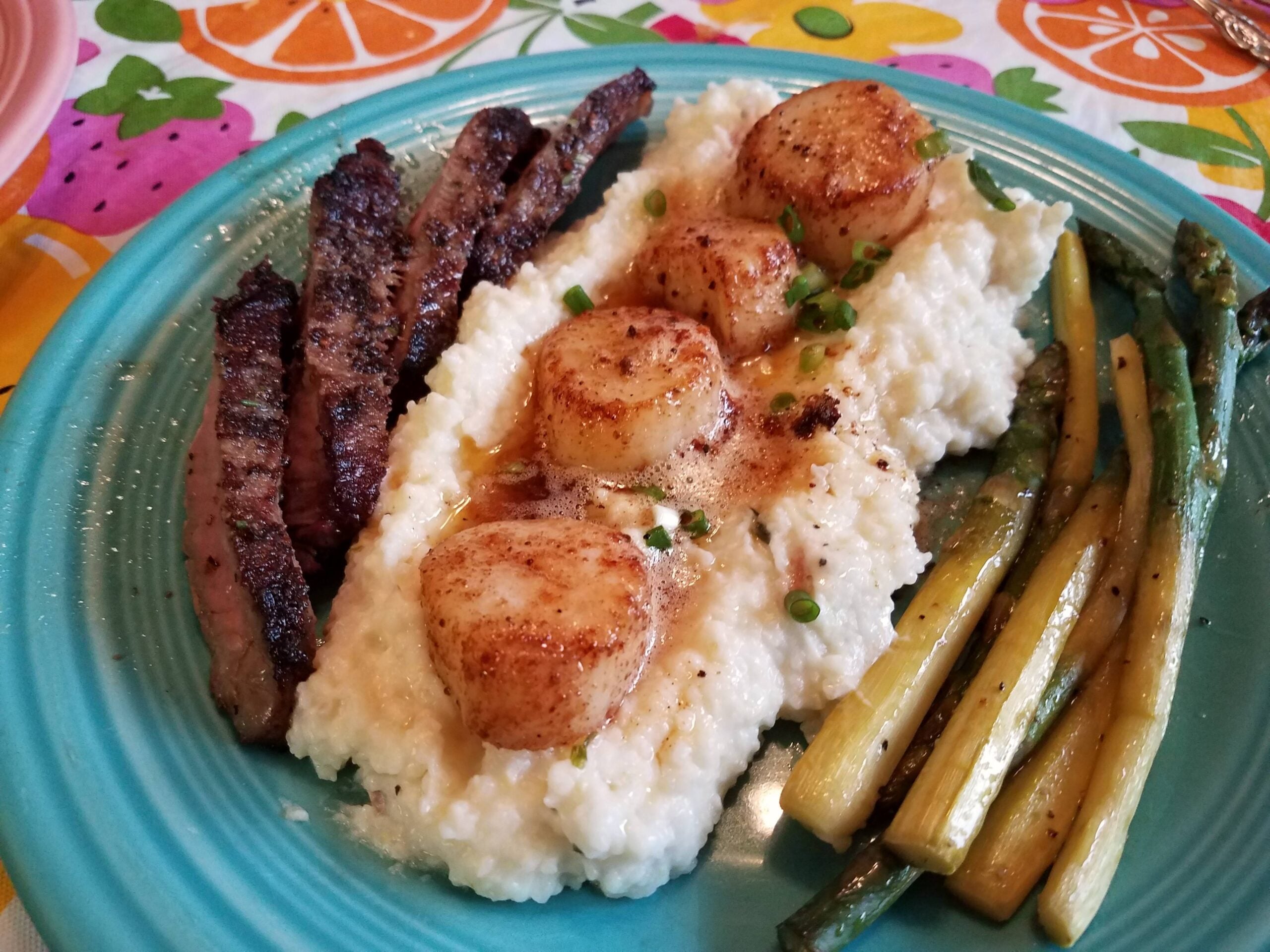 A plate with grilled steak, seared scallops on creamy grits topped with chives, and roasted asparagus, served on a turquoise dish with a colorful tablecloth underneath.