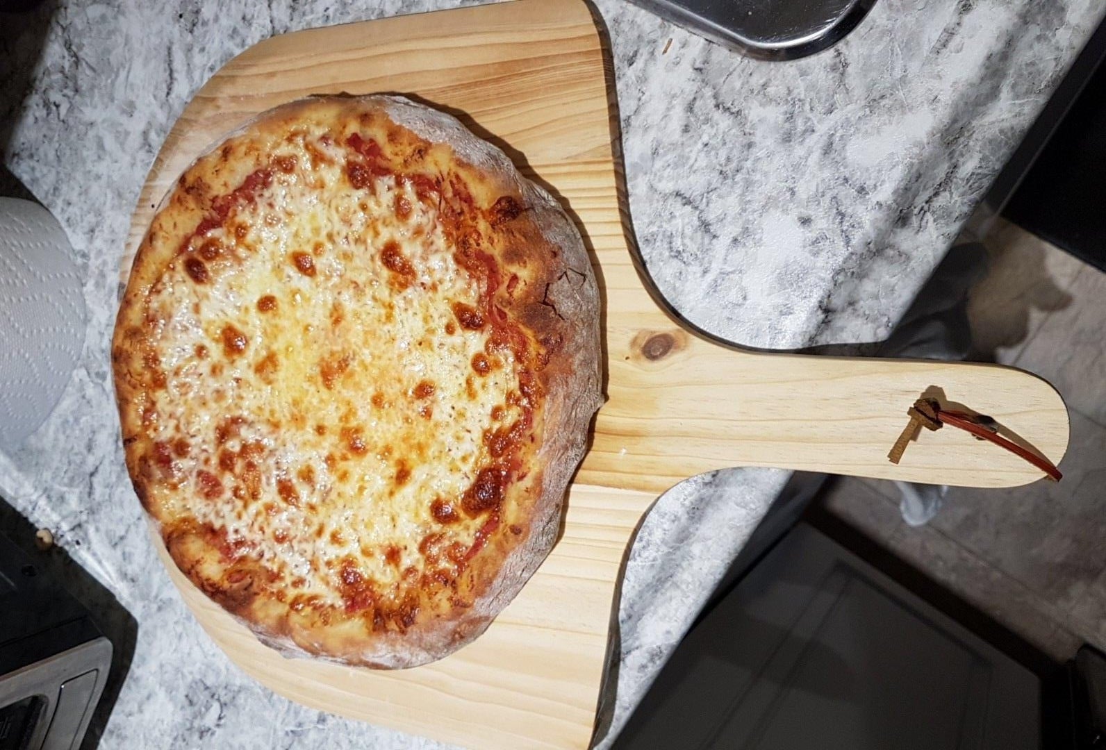 A freshly baked cheese pizza sits on a wooden pizza peel, atop a marble countertop. The pizza has a golden, bubbly cheese topping and a thick crust.