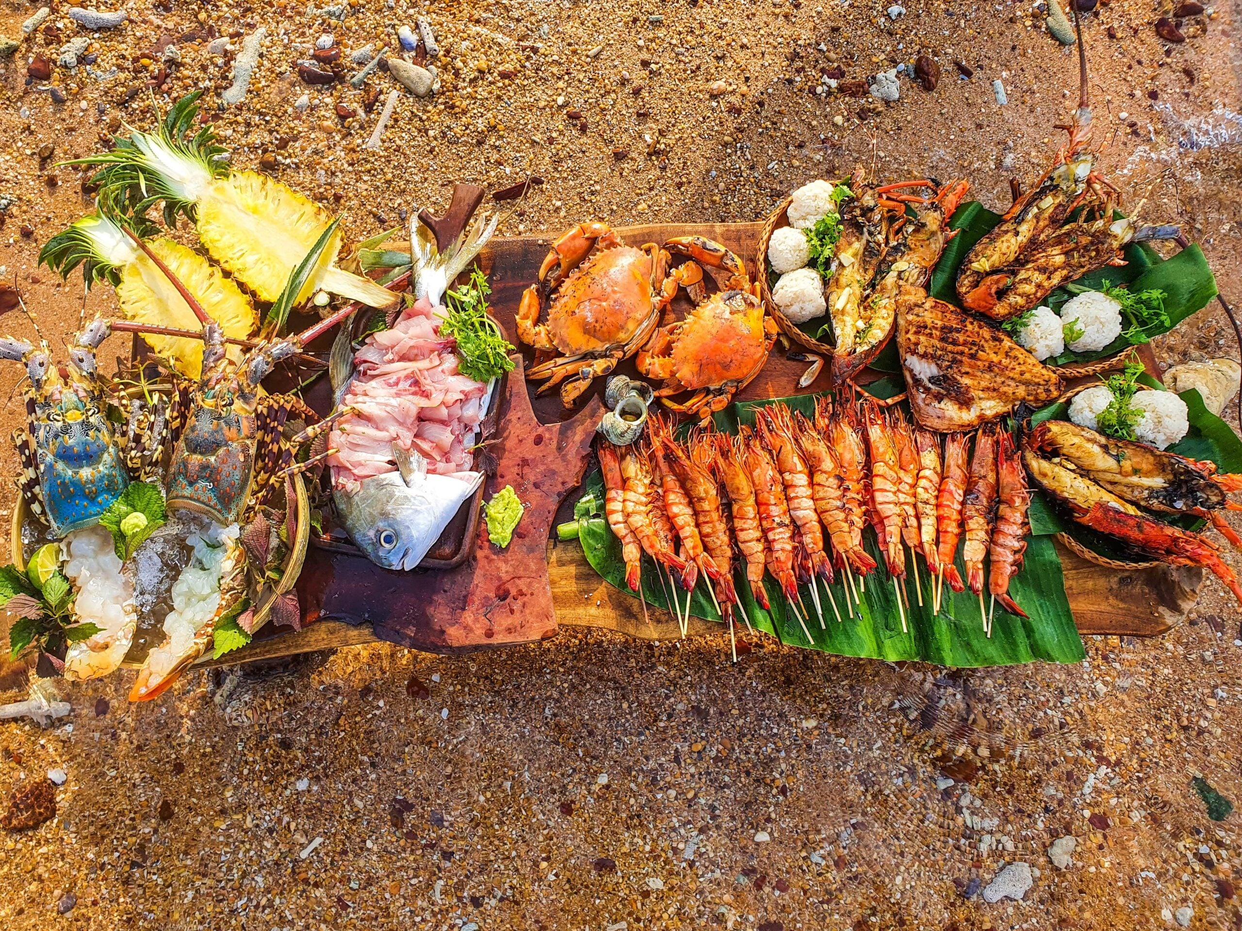 A colorful seafood feast on a wooden platter with grilled shrimp, crabs, fish, oysters, pineapple slices, and rice balls, arranged on green leaves on a sandy beach.
