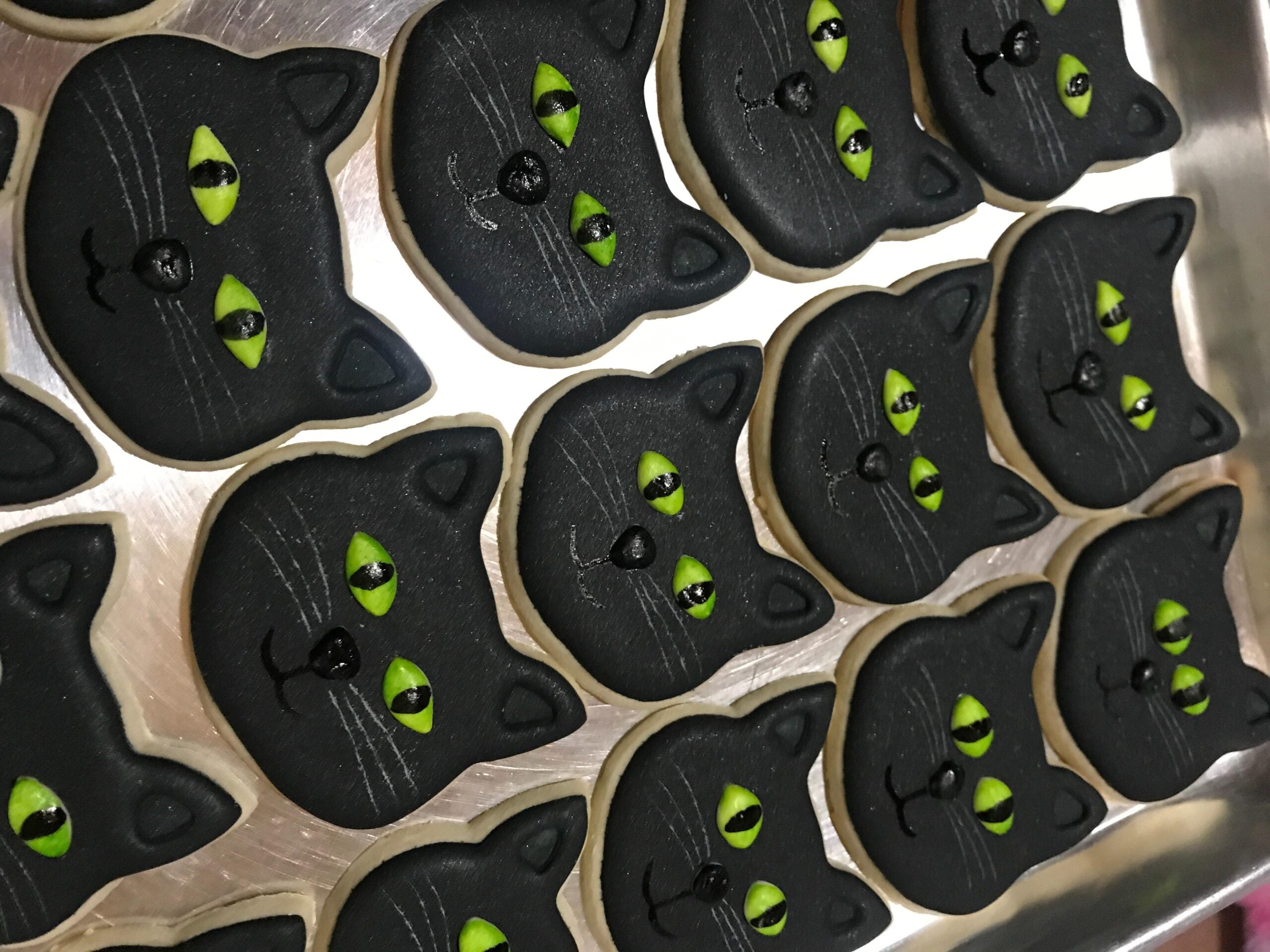 A tray of cookies decorated as black cat faces with green eyes and white whiskers, neatly arranged in rows on a metal surface.
