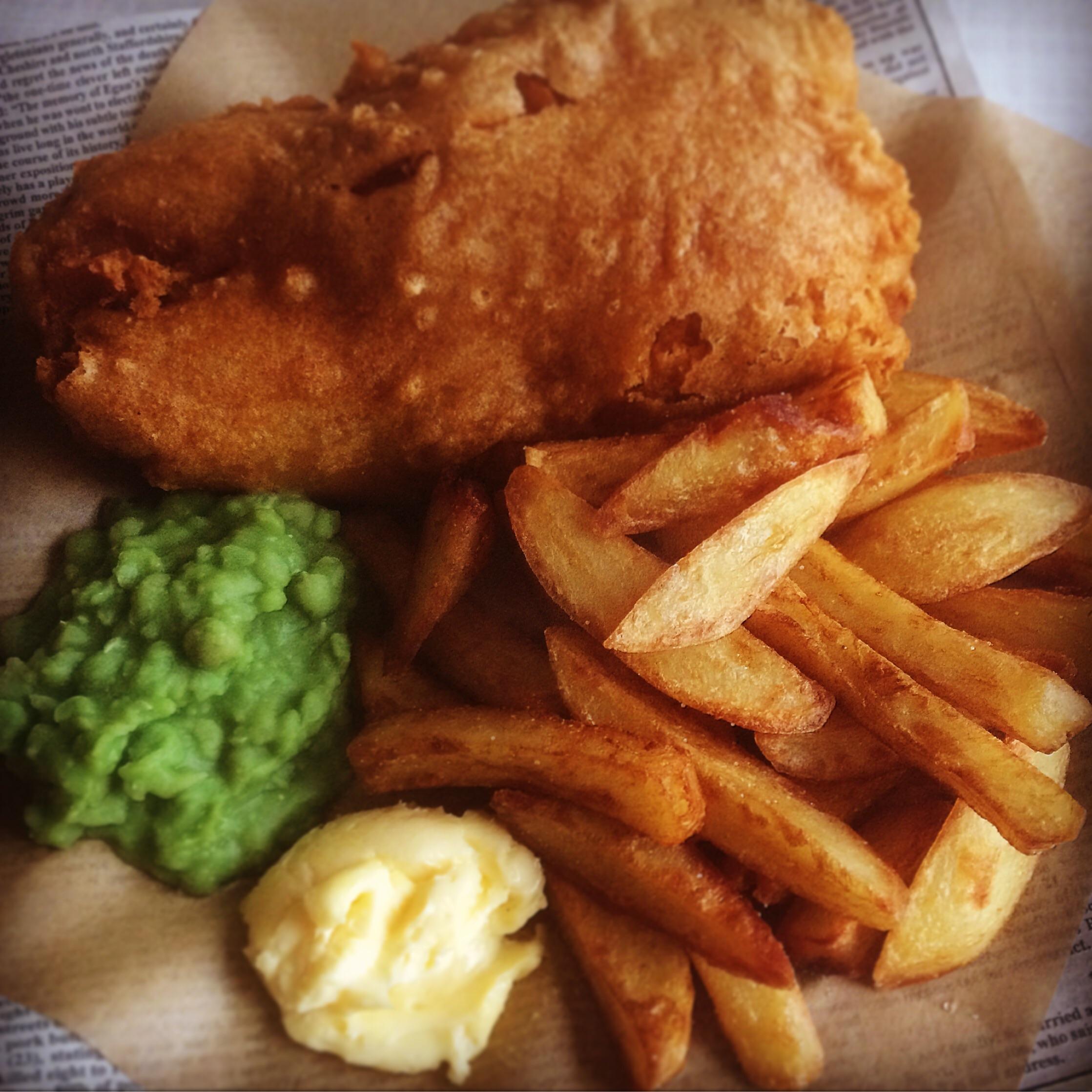 A serving of battered fried fish, thick-cut fries, mushy peas, and a dollop of tartar sauce on brown paper with newspaper print.