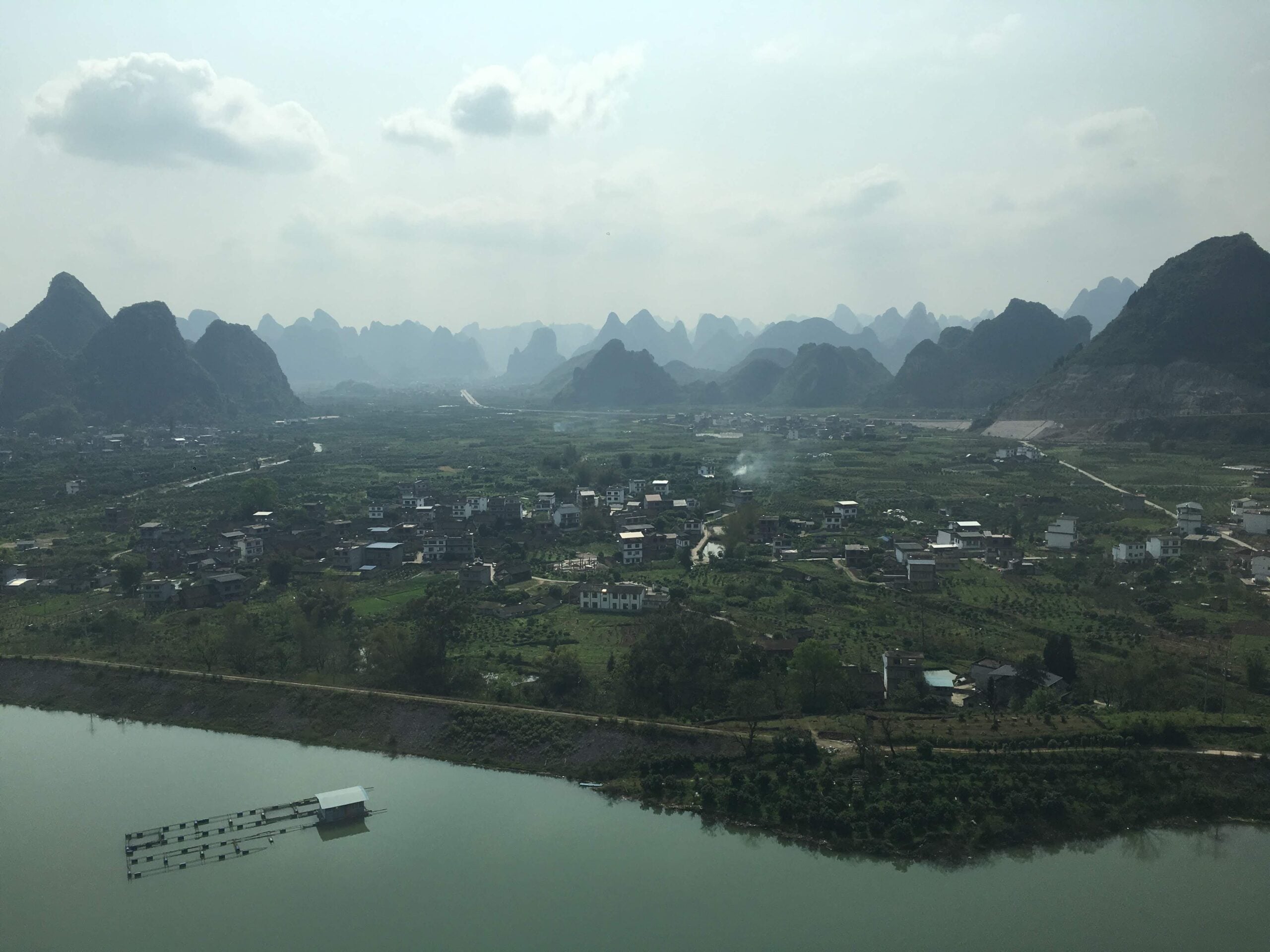 A river runs along a lush village with scattered houses, backed by dramatic, misty karst mountains under a cloudy sky. A floating dock sits on the river’s edge.