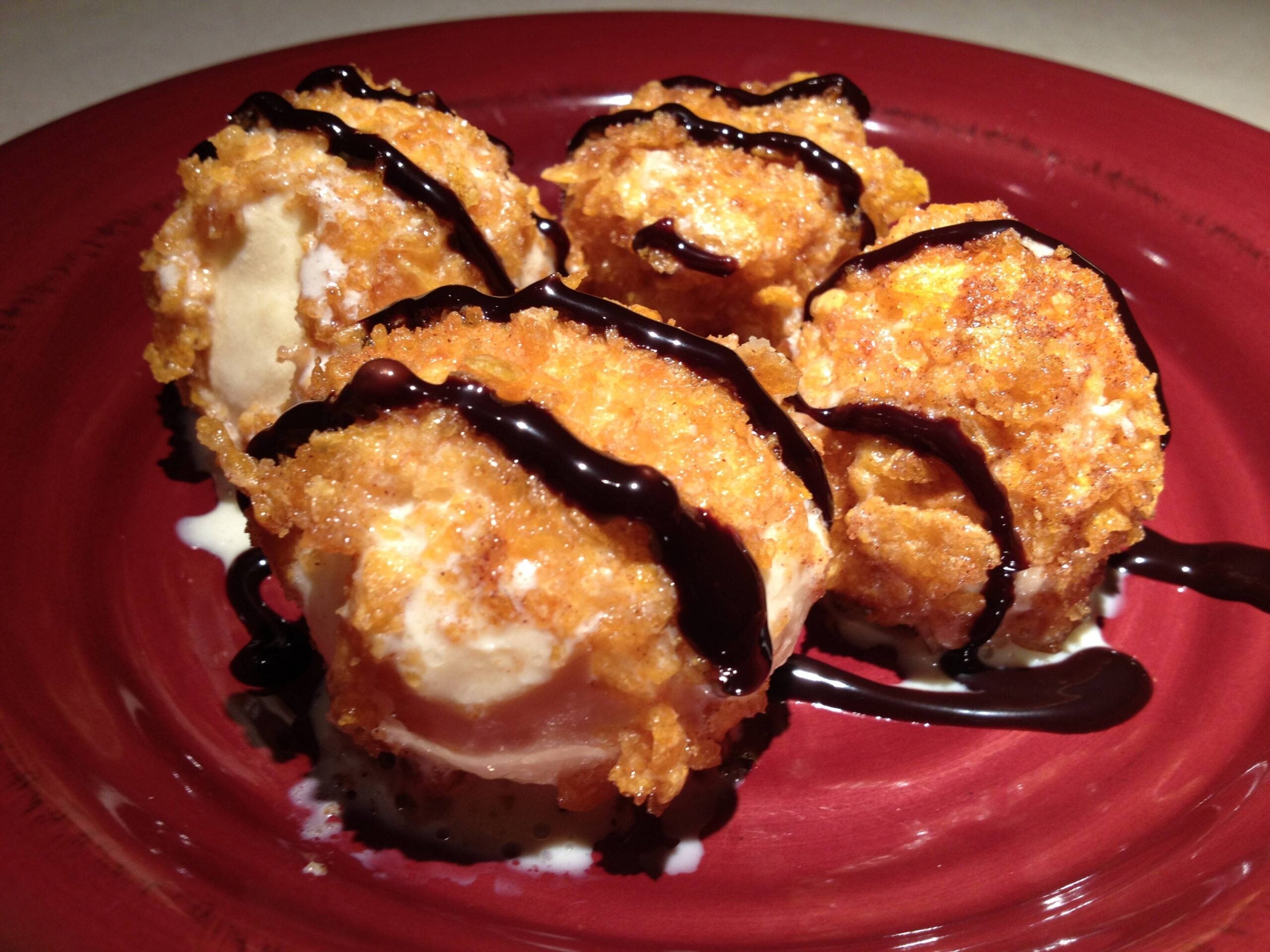 Four scoops of fried ice cream with crispy golden coating, drizzled with chocolate sauce, served on a red plate.
