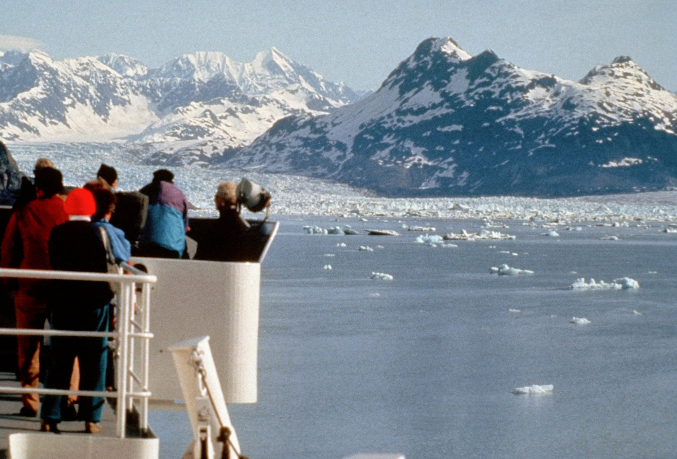 People stand on a ship’s deck, enjoying trips where getting there means breathtaking views of icy water and snow-covered mountains dotted with icebergs under a clear sky.