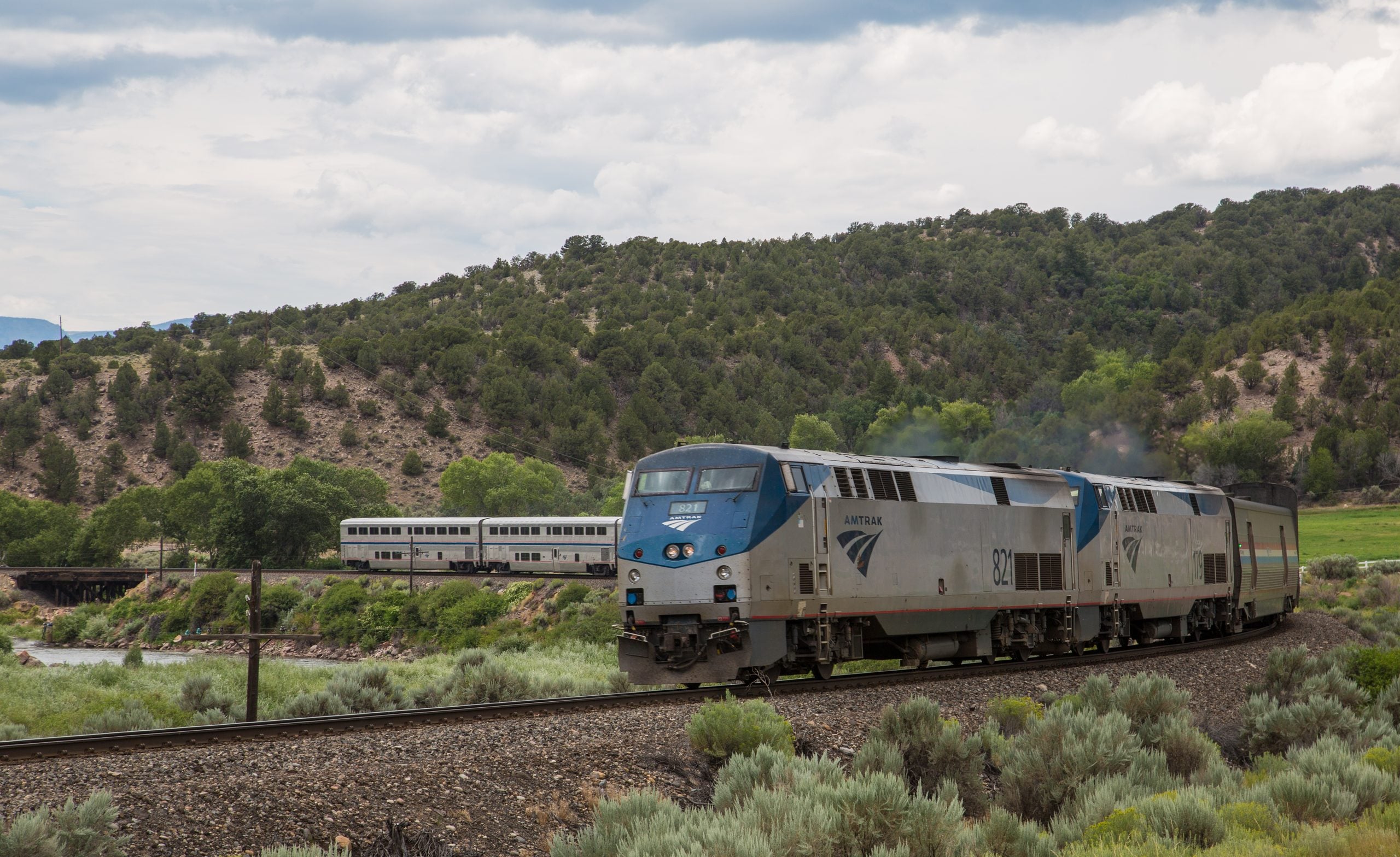 An Amtrak train with locomotives numbered 821 pulls passenger cars through a rural, hilly landscape, perfect for trips where getting there is part of the adventure, with sagebrush and trees under a cloudy sky.