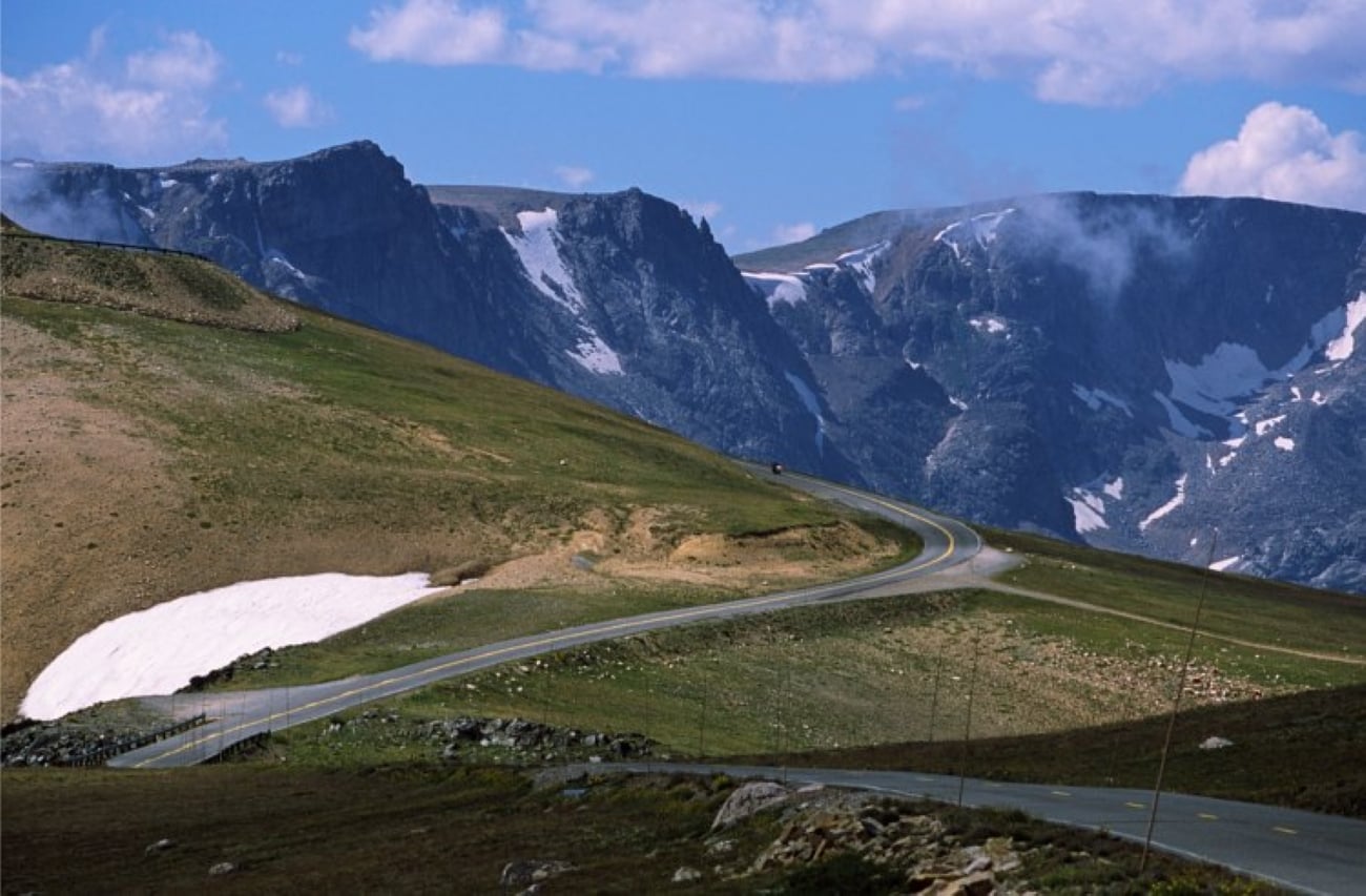 A winding road curves through grassy hills with patches of snow, perfect for trips where getting there is part of the adventure, leading toward rugged mountains under a partly cloudy blue sky.