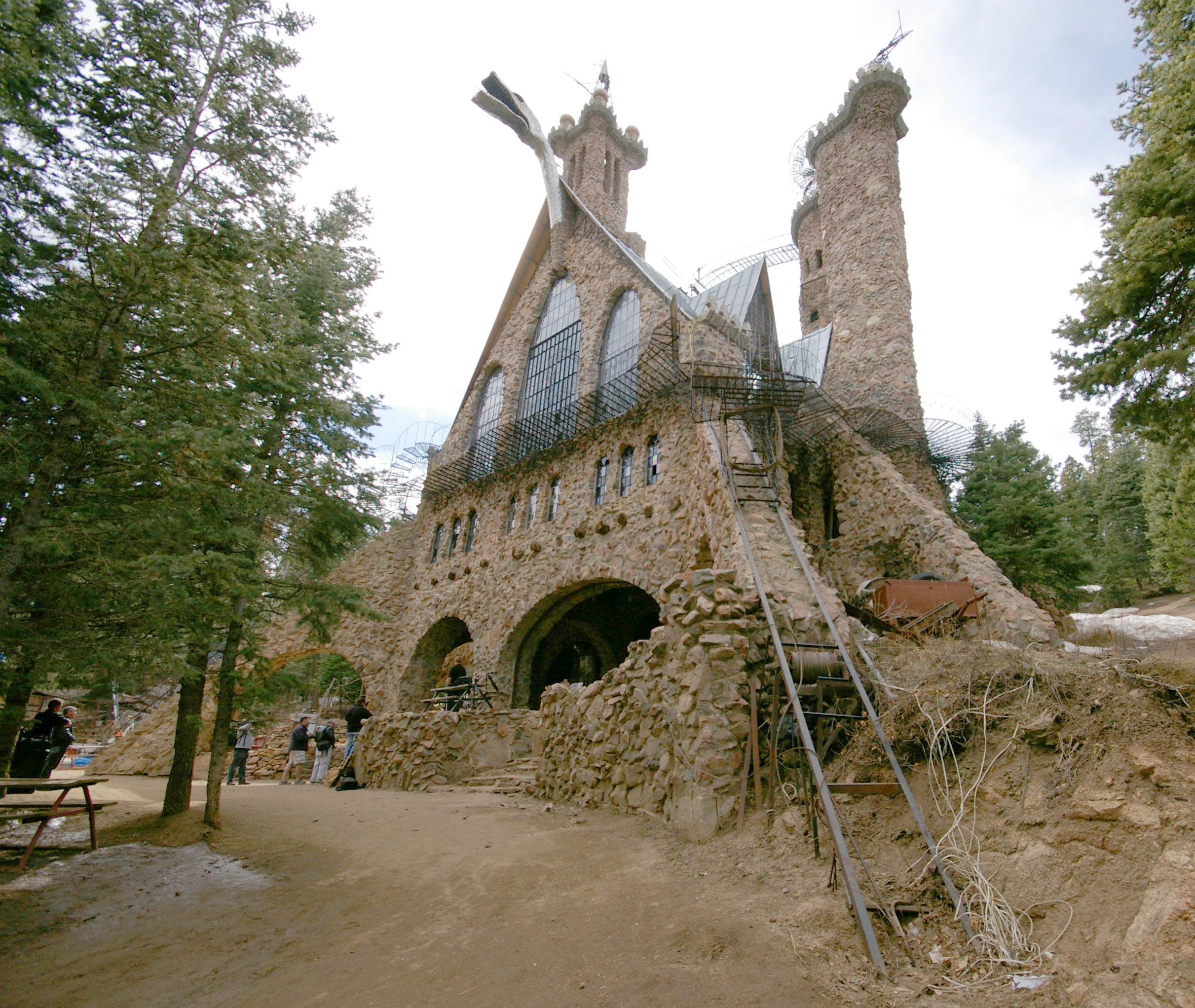 Stone castle structure with tall towers, arched entrances, and metal framework, surrounded by pine trees. Reminiscent of the grand castles of old, a dirt path and wooden ladders lead to the entrance. People are gathered near the building.