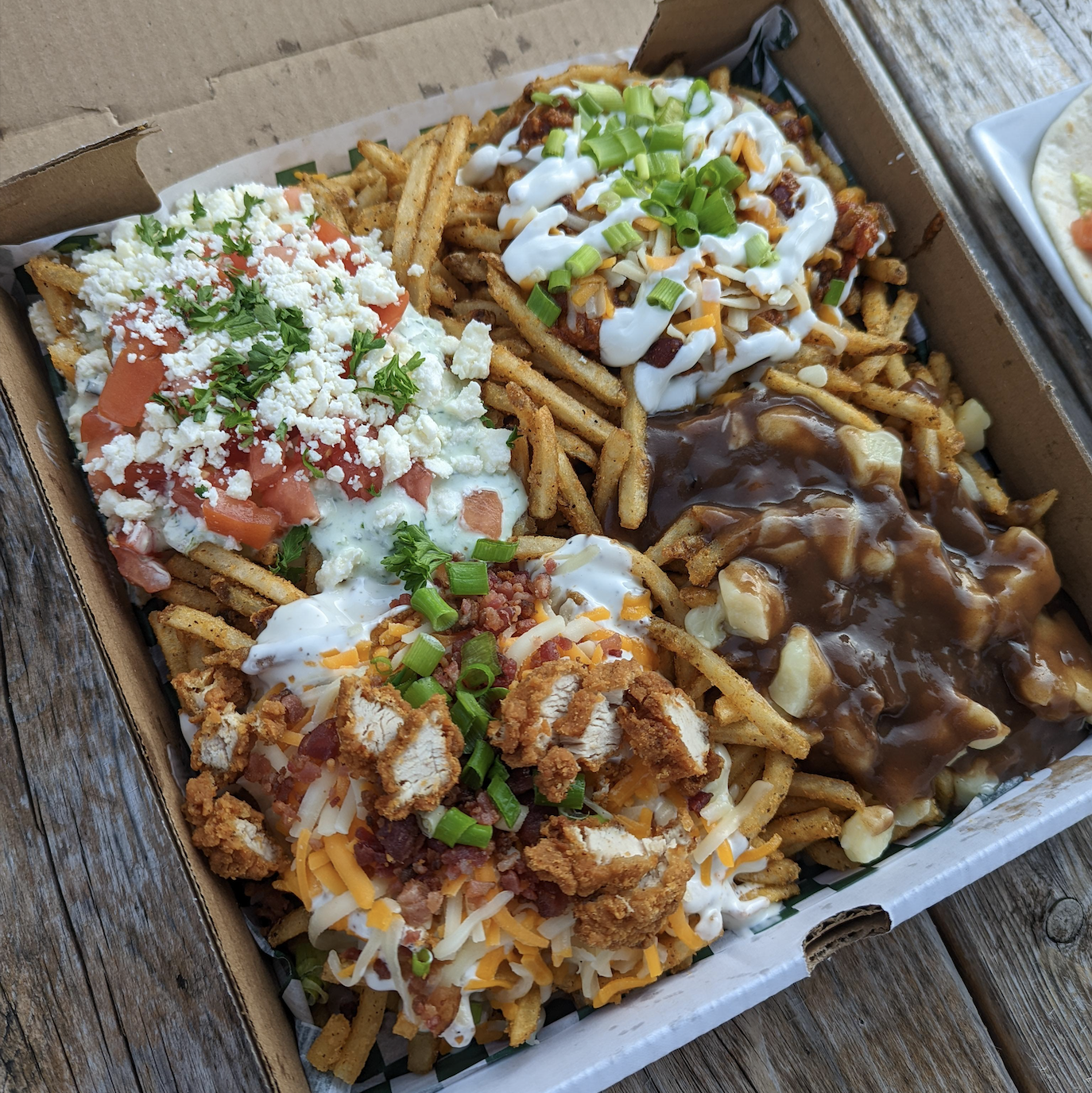 A box of loaded fries with four sections, each topped differently: chili, cheese, green onions, sour cream, gravy with cheese curds, diced tomatoes, crumbled cheese, and fried chicken pieces, on a wooden table.