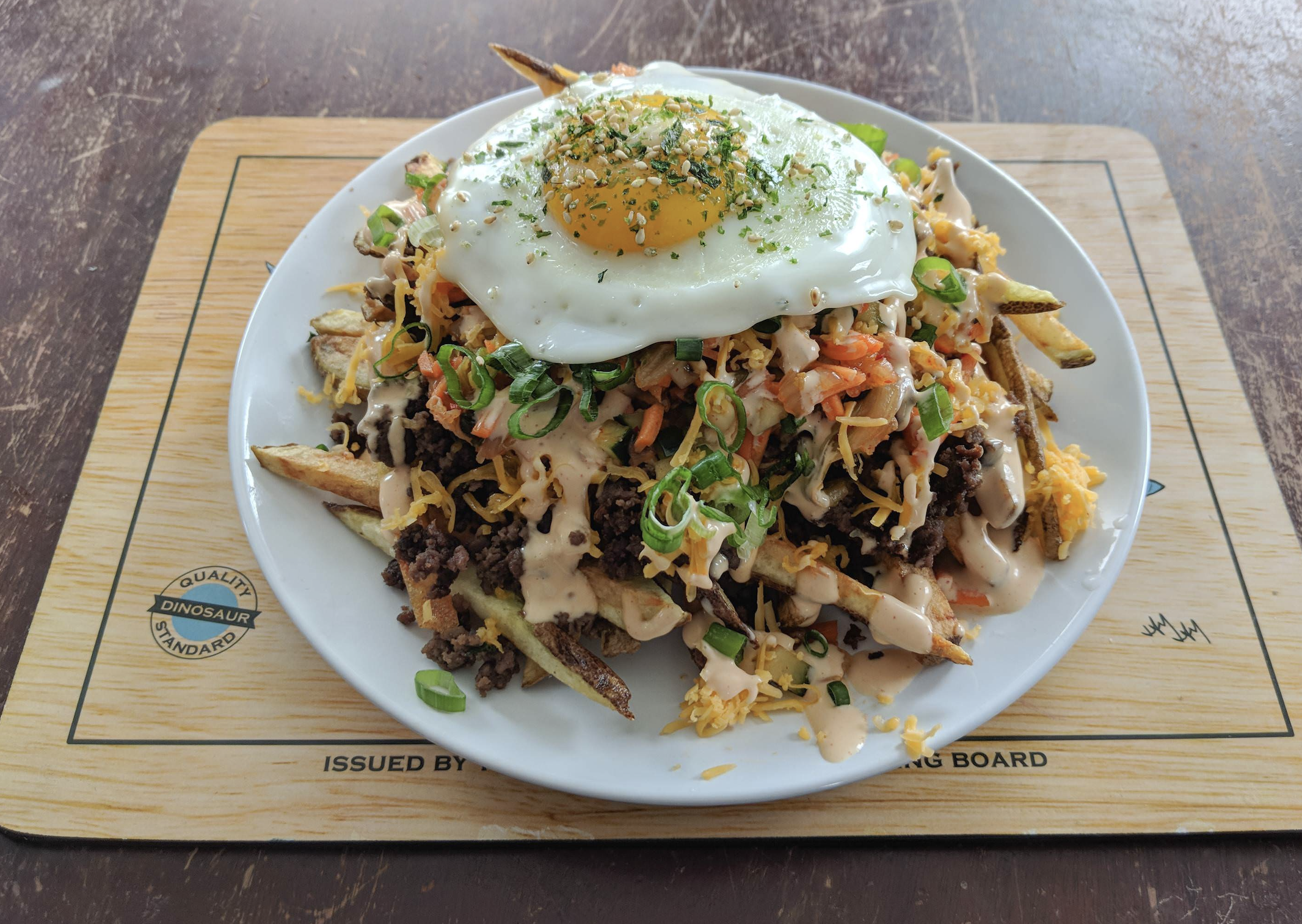 A plate of loaded fries topped with minced meat, cheese, chopped green onions, drizzled sauce, kimchi, and a sunny-side-up egg, served on a white plate over a wooden board.