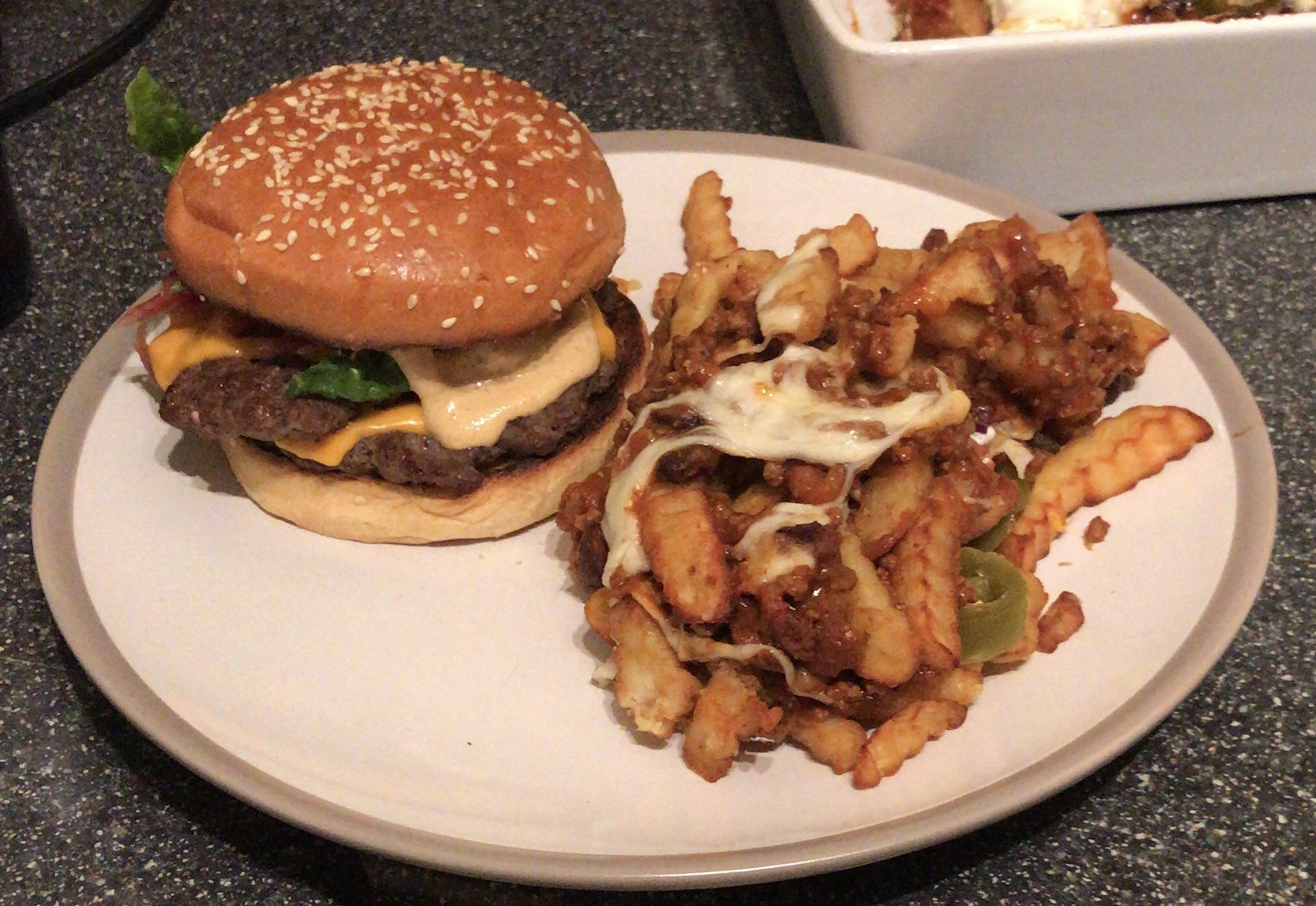 A cheeseburger with lettuce and sauce sits next to a serving of crinkle-cut fries topped with melted cheese and chili on a white plate.