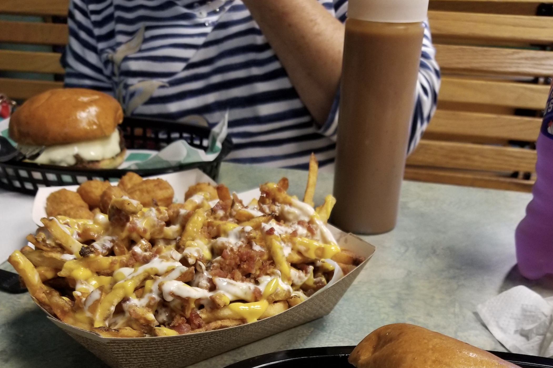A tray of loaded fries with cheese, bacon, and sauce is on a table, with a burger, tater tots, a bottle of sauce, and a person in a striped shirt in the background.