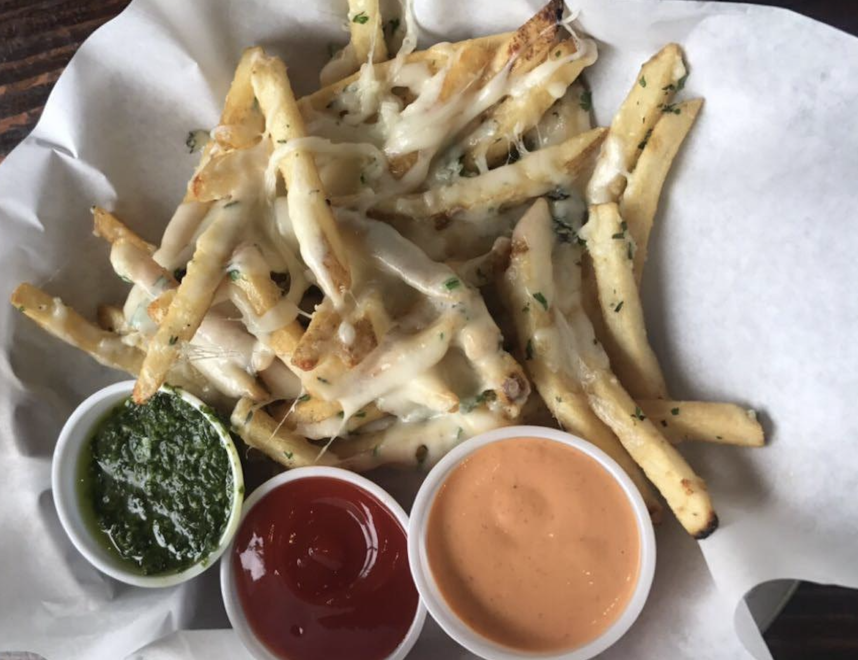 A basket of cheesy garlic fries is served with three dipping sauces: green pesto, red ketchup, and orange chipotle mayo, on white parchment paper.