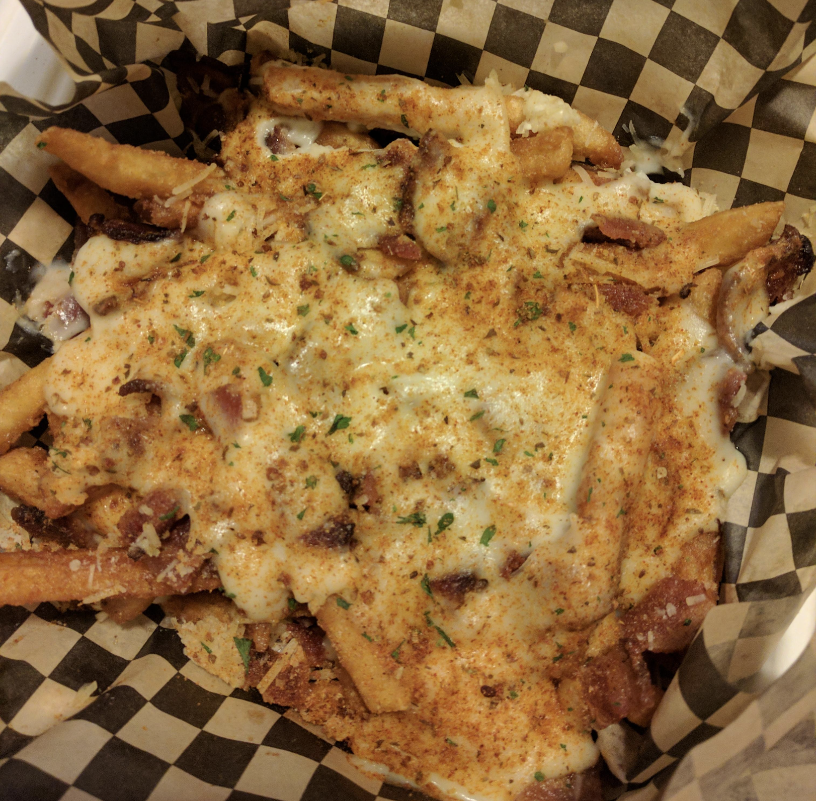 French fries topped with melted cheese sauce, bacon bits, and seasoned bread crumbs, served in a basket lined with black and white checkered paper.