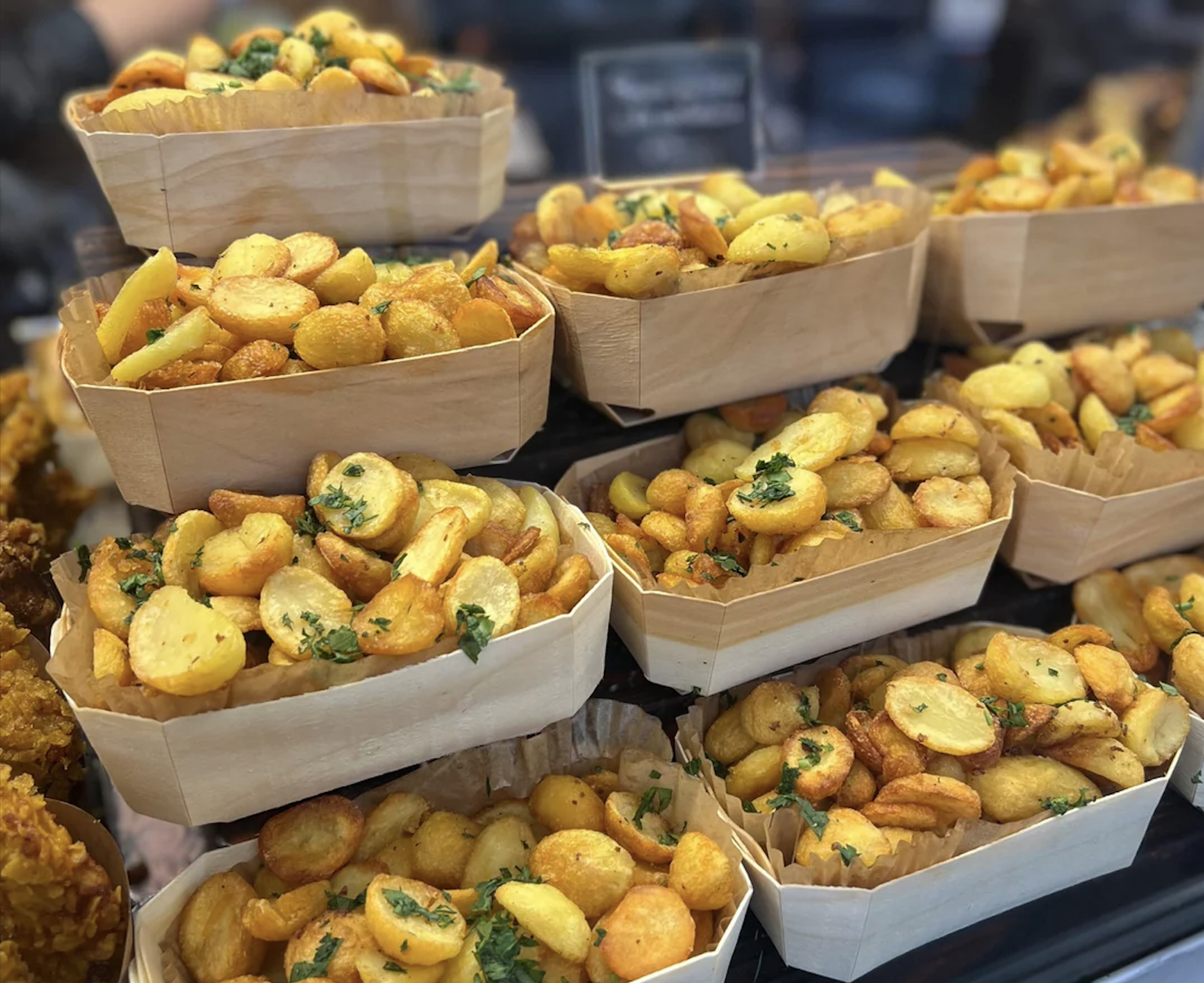 Several wooden trays are filled with golden, seasoned roasted potatoes, garnished with chopped herbs, stacked in a display at what appears to be a market or food stall.