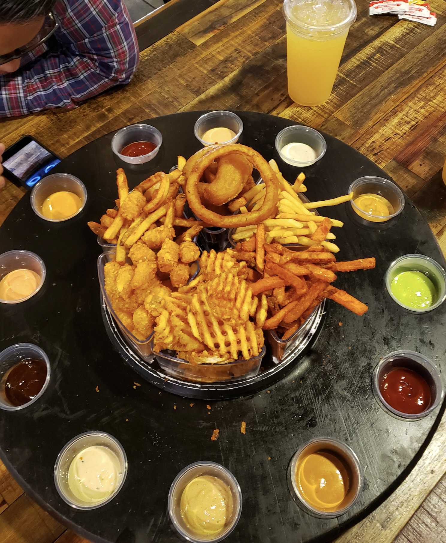 A platter of fries, onion rings, and fried bites sits on a table, surrounded by various dipping sauces in small cups. A drink and a person using a phone are visible in the background.