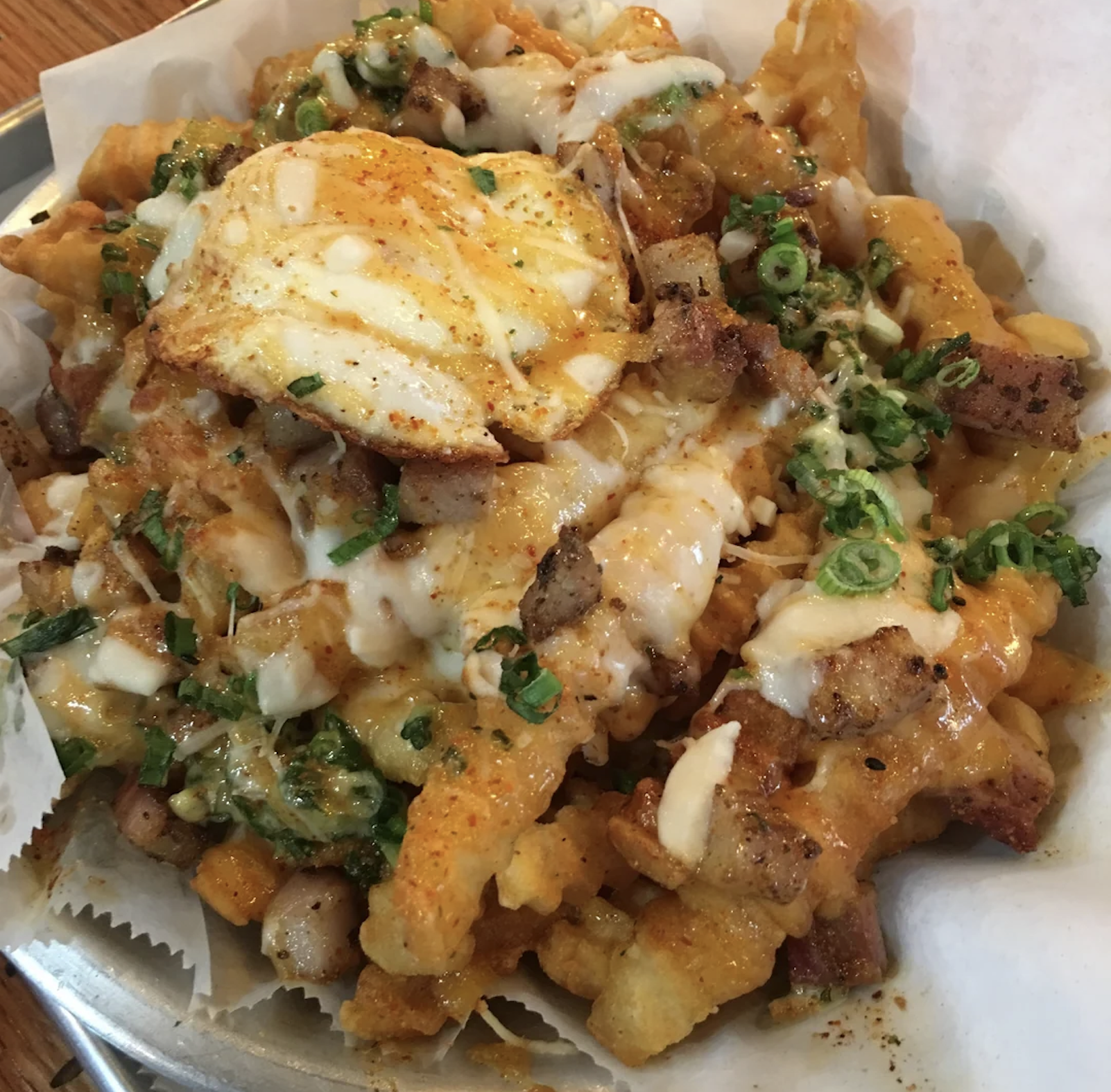 A close-up of loaded crinkle-cut fries topped with melted cheese, green onions, diced meat, spices, and a fried egg served in a paper-lined basket.