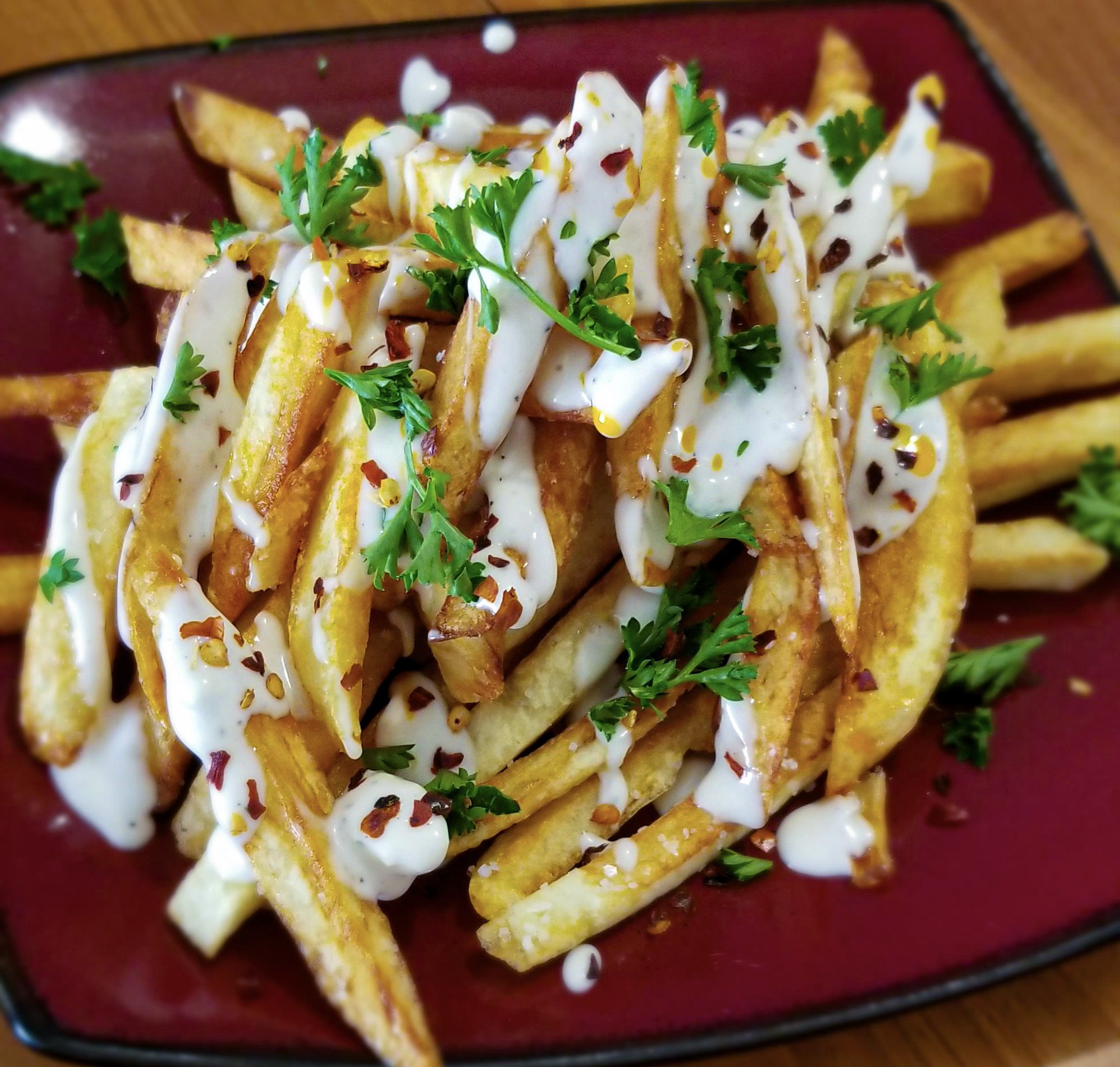 A plate of golden French fries topped with creamy white sauce, chopped parsley, and red pepper flakes, served on a red square dish.