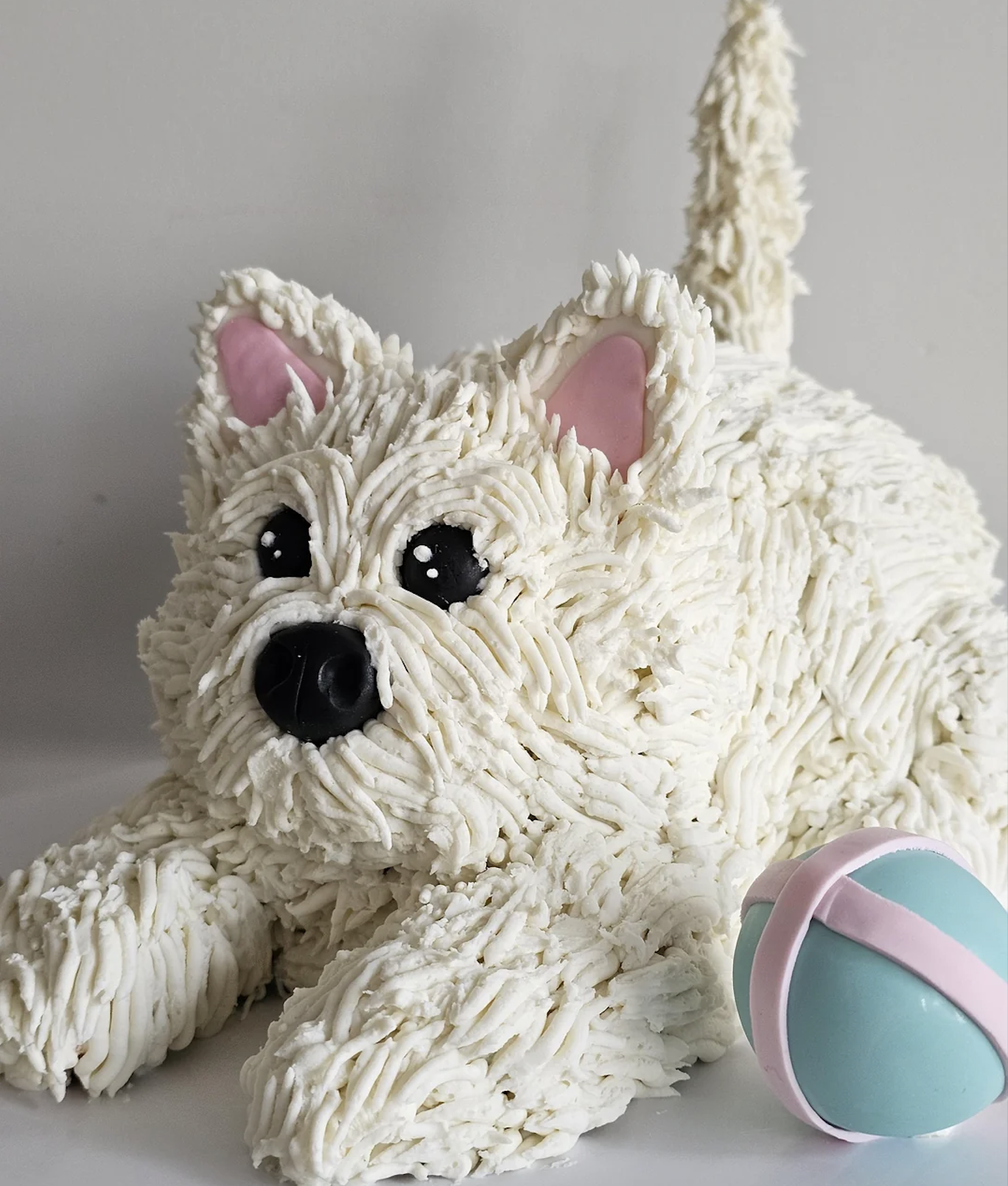 A cake shaped like a fluffy white dog with pink ears and a playful expression lies next to a pastel blue and pink ball.