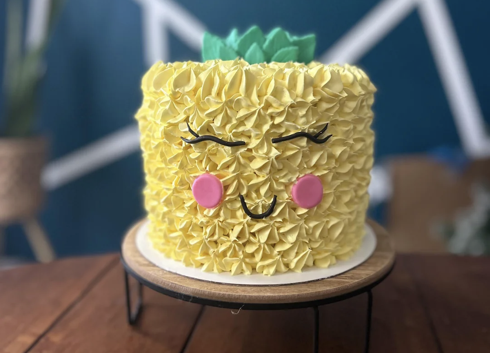 A round cake decorated to look like a cute, smiling pineapple with yellow textured frosting, pink cheeks, closed eyes, and green leaves on top, set on a wooden stand.