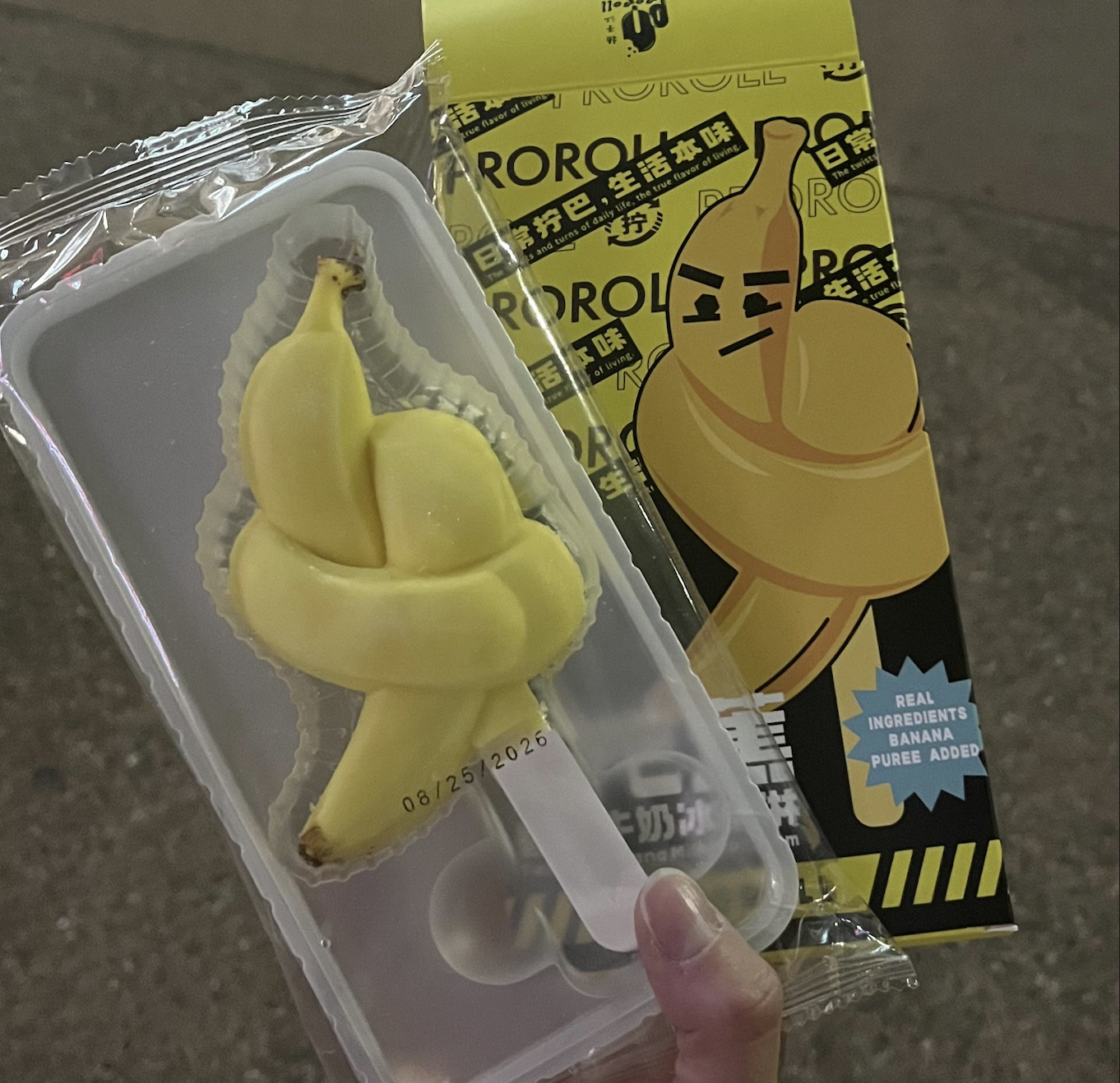 A hand holds a yellow ice cream shaped like a twisted banana, next to its packaging featuring a cartoon banana with a serious face. The label mentions real banana puree added.