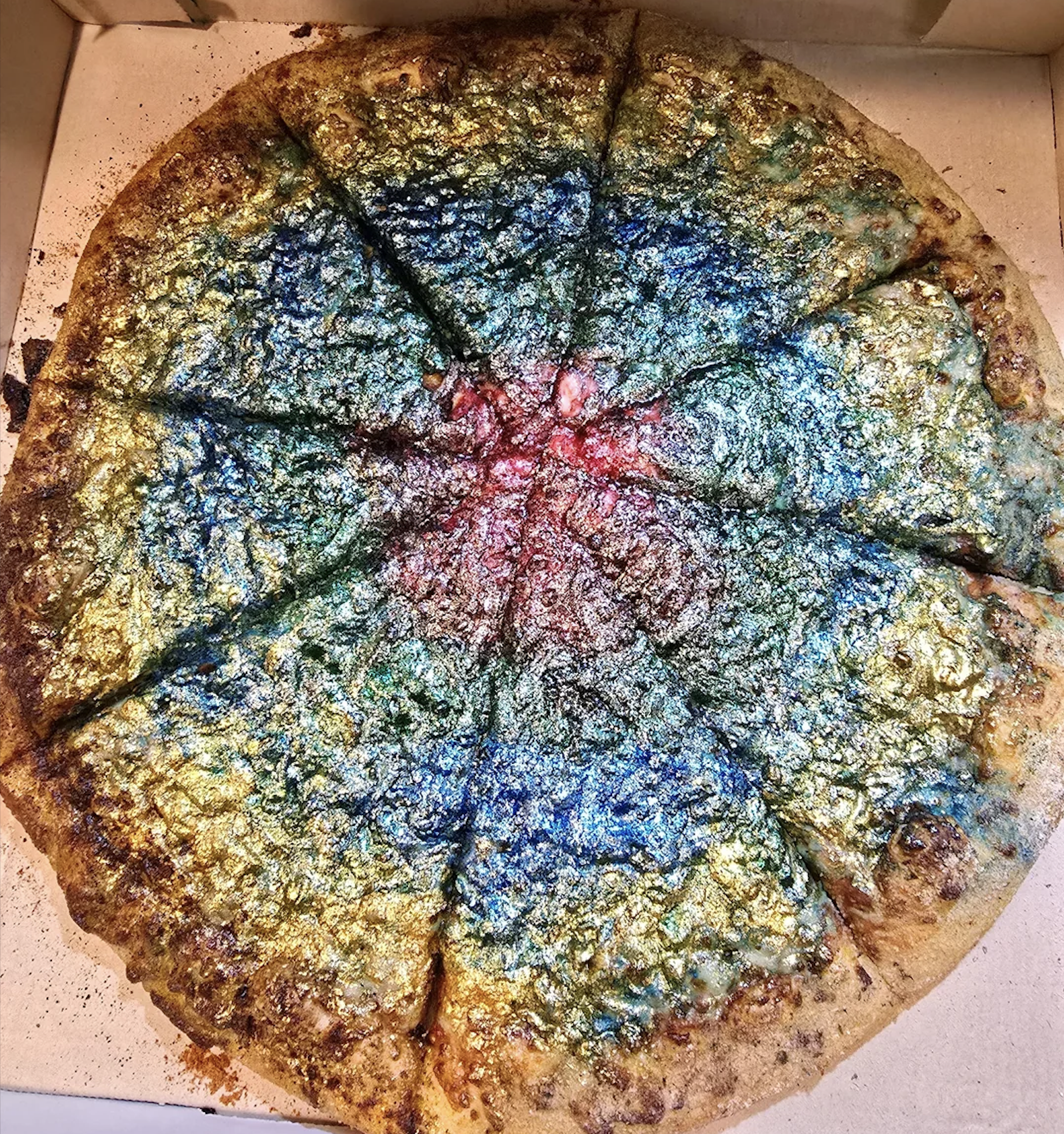 A whole pizza sliced into eight pieces, topped with shiny, colorful, metallic-looking glitter in blue, green, yellow, and red hues. The vibrant glitter covers the entire surface evenly.