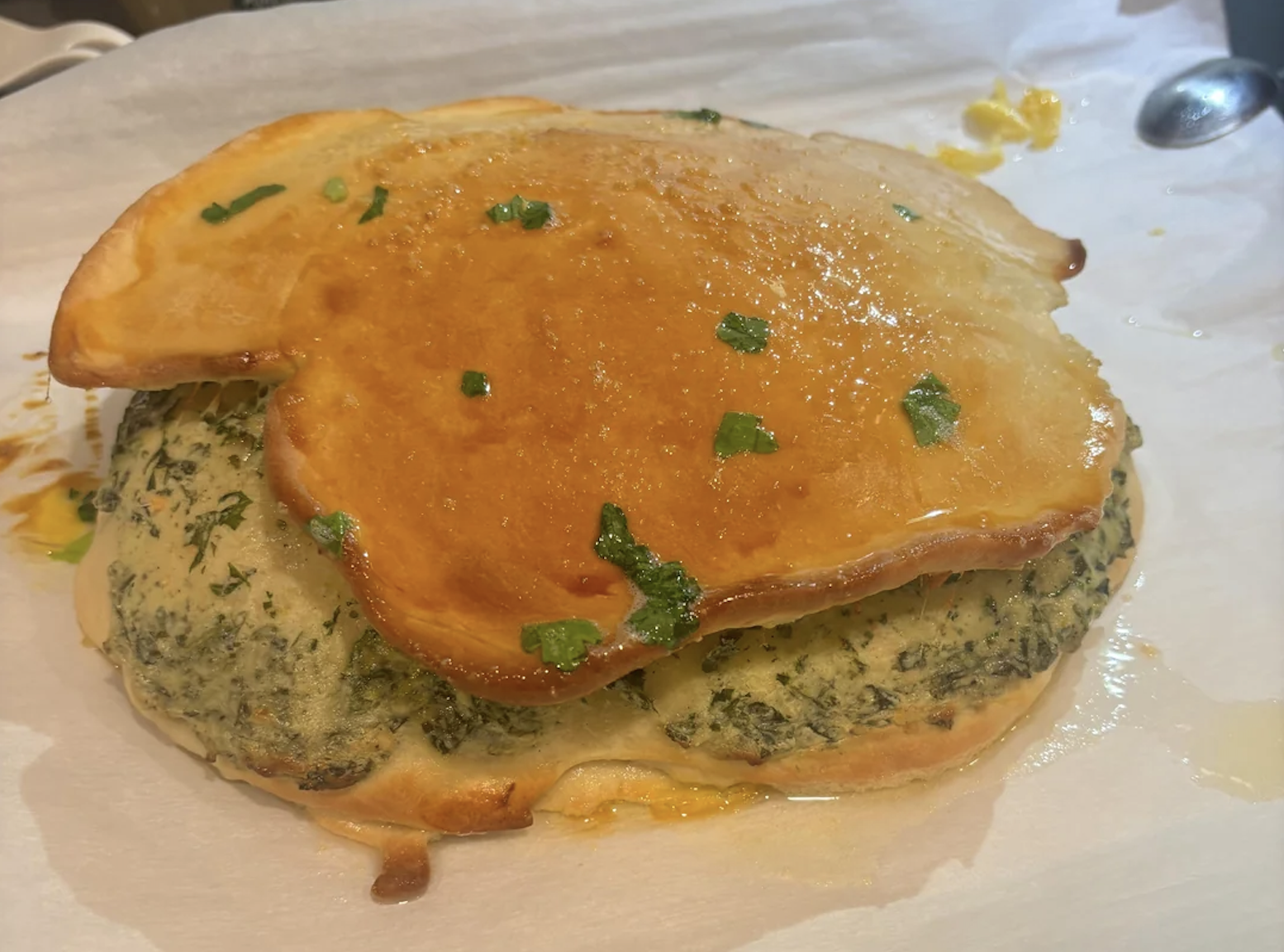 A baked dish with a golden, glazed crust on top of a green herb-filled base, resting on parchment paper with some melted butter and herbs visible.