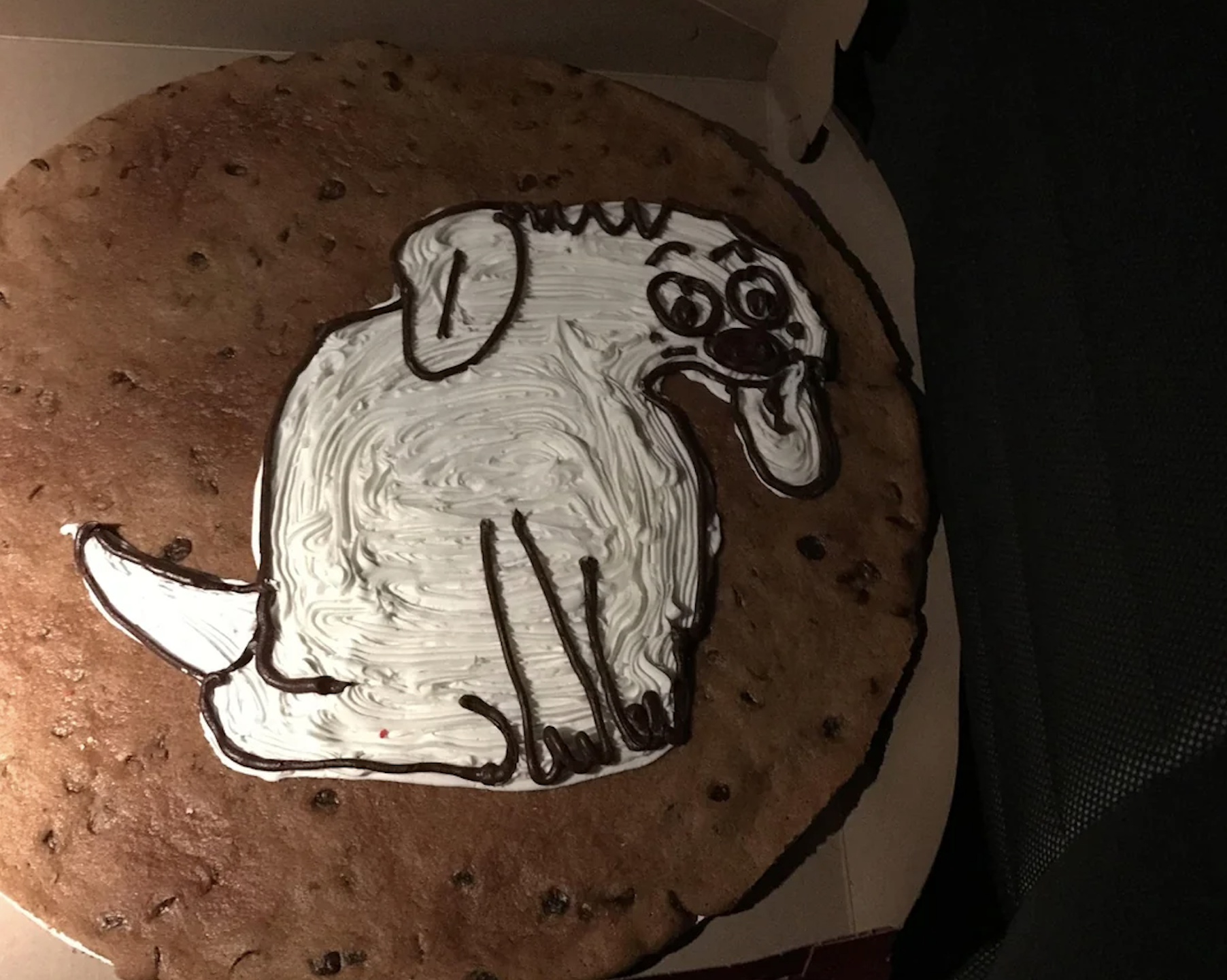 A large chocolate chip cookie with a cartoon dog drawn in white and brown icing, sitting with its tongue out, inside a pizza-style box.