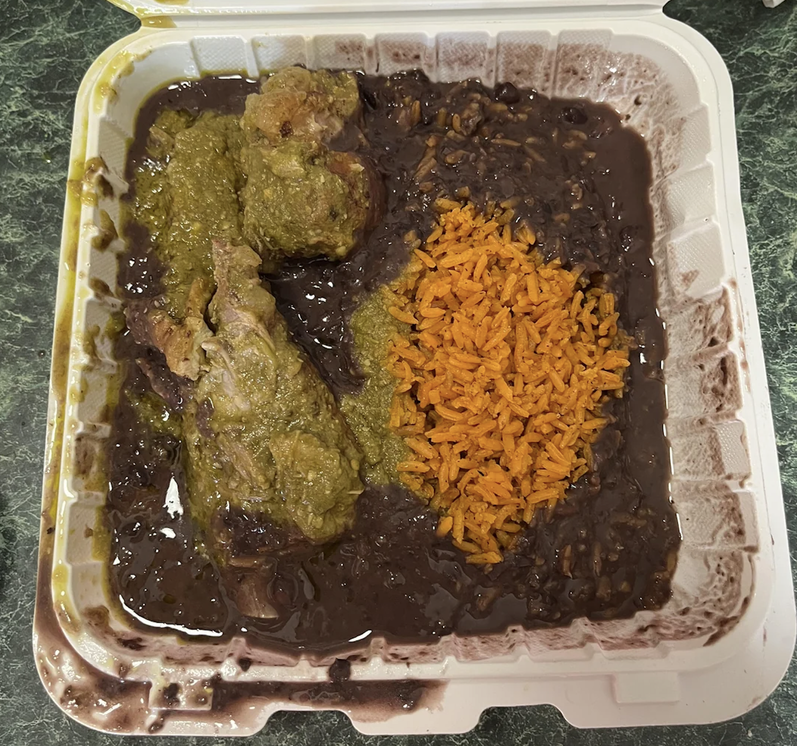 Takeout container with pork in green sauce, orange rice, and black beans, with some sauce spilling into the sections, placed on a green countertop.
