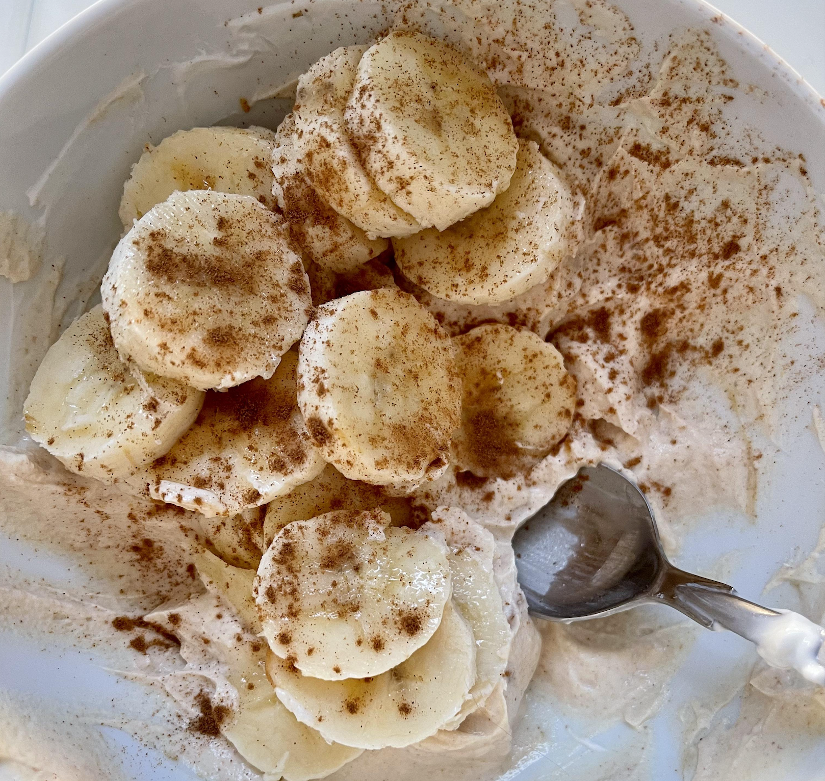Sliced bananas topped with cinnamon sit on a creamy mixture in a white bowl. A spoon rests in the bowl, partially covered by the creamy base and banana slices.