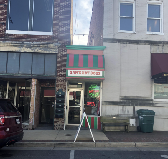 A narrow storefront with a red and green striped awning reads &ldquo;Sam&rsquo;s Hot Dogs.&rdquo; A &ldquo;HOT DOGS&rdquo; neon sign and an &ldquo;OPEN&rdquo; sign are in the window. A sidewalk sign stands outside. The building is between two larger structures.