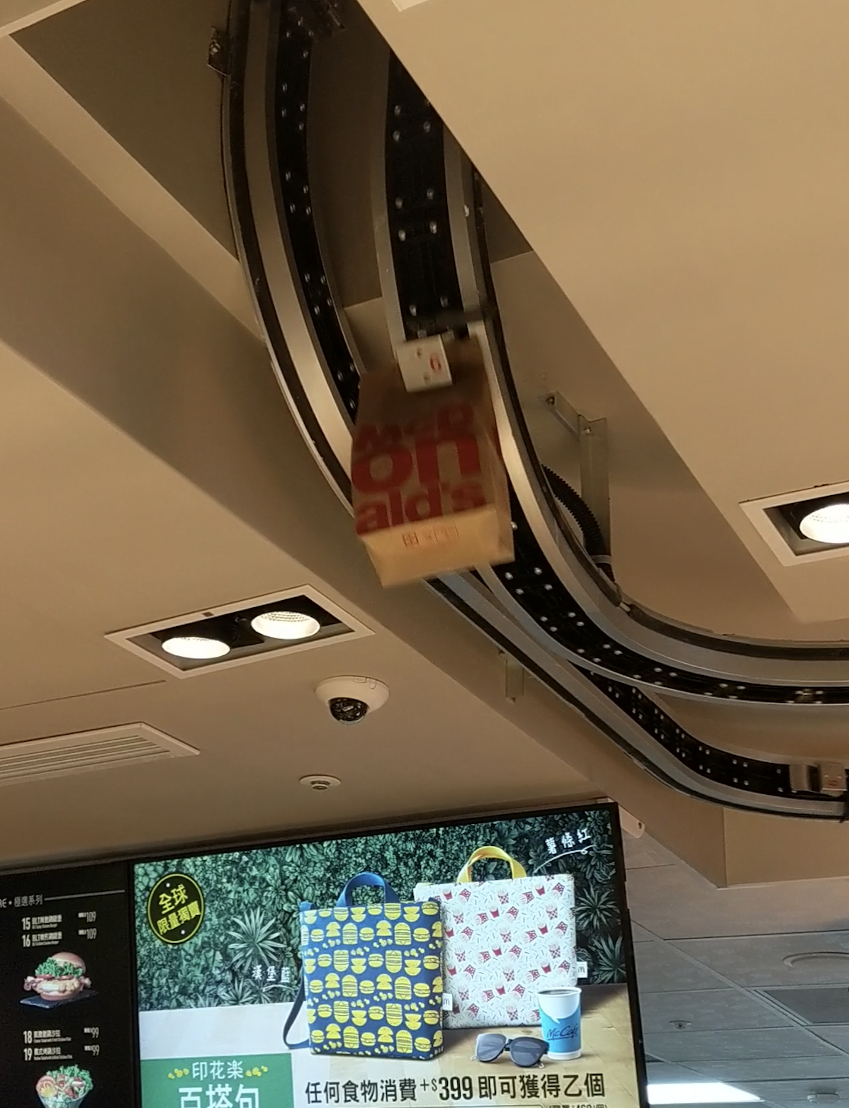 A paper bag moves along a ceiling conveyor system above a fast-food restaurant counter, with a display screen showing promotion and colorful bags below.