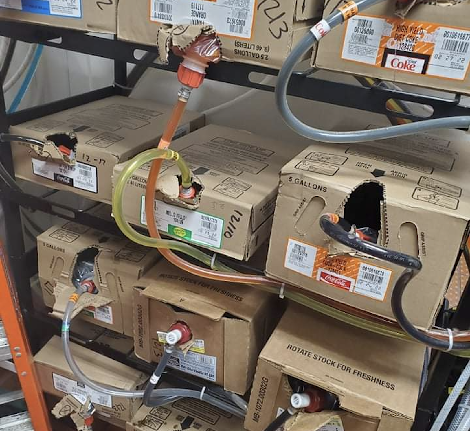 Several syrup boxes with tubes connected to dispensers are stacked on metal shelves. Each box is labeled with beverage names like Diet Coke and Sprite. Some boxes have holes for tube access.