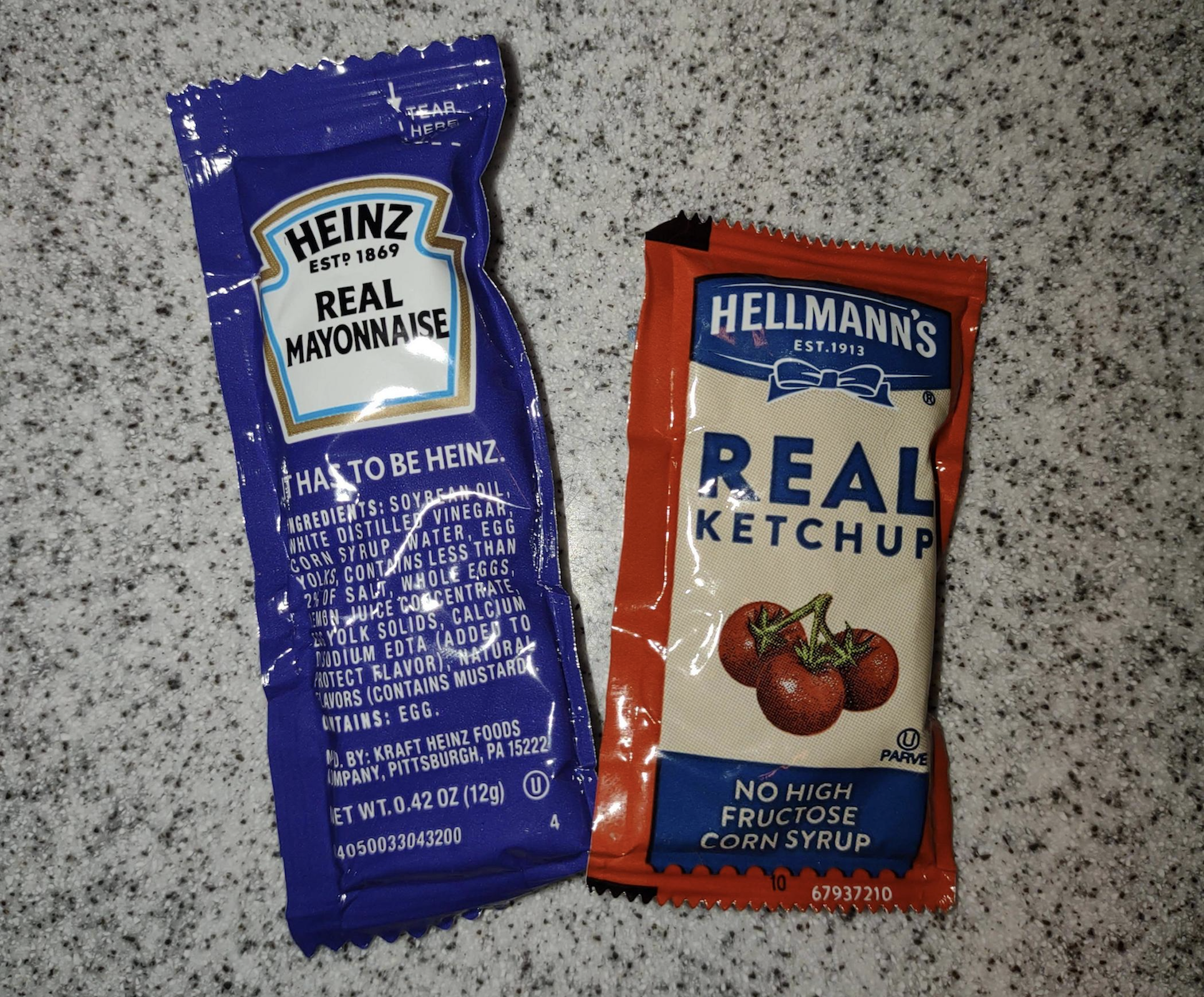 Two condiment packets on a counter: one Heinz Real Mayonnaise in a blue packet and one Hellmann’s Real Ketchup in a red packet, both placed side by side on a speckled surface.