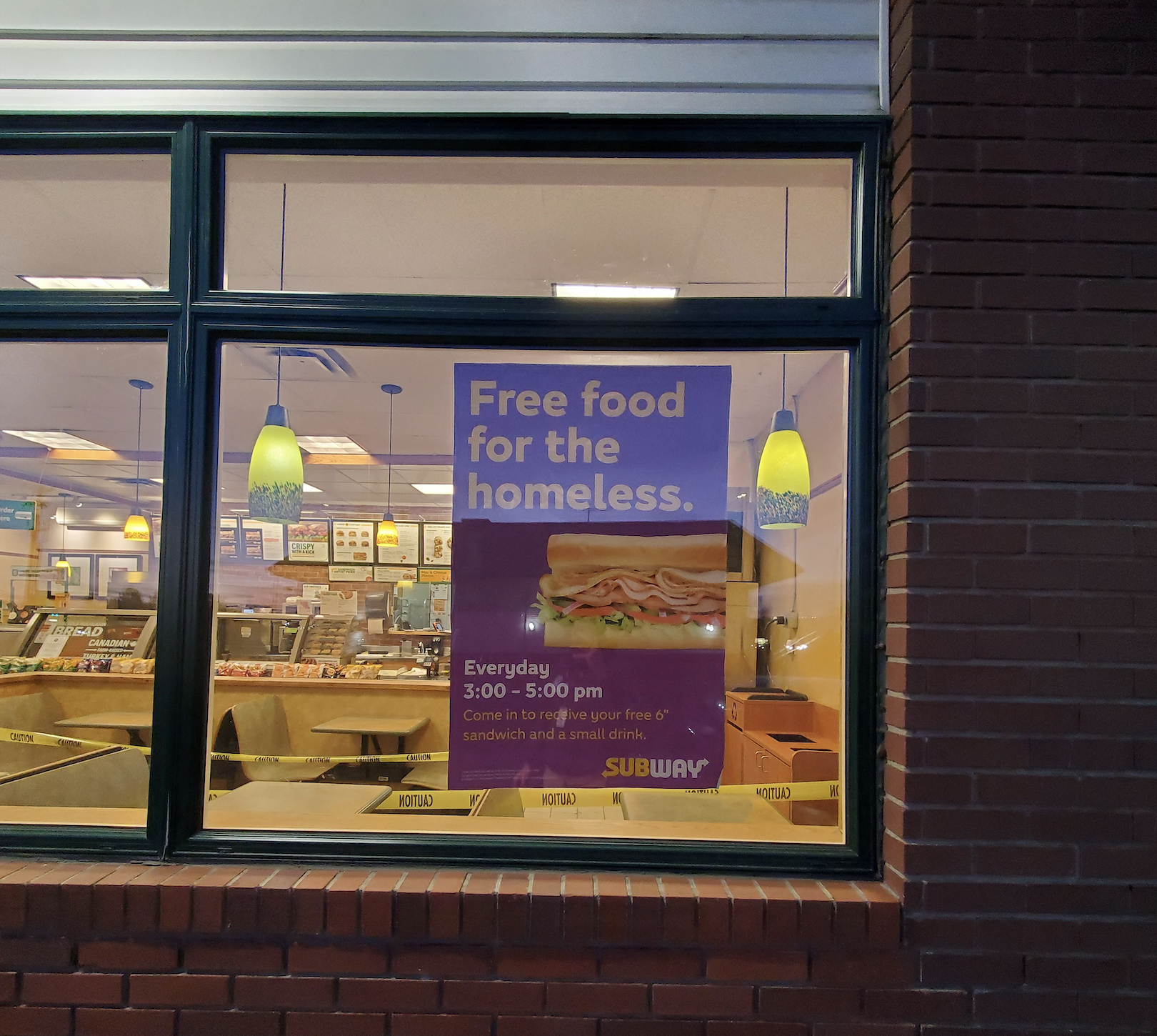 A Subway restaurant window displays a sign offering free food for the homeless, with a sandwich image and details about the promotion running daily from 3:00 to 5:00 pm.