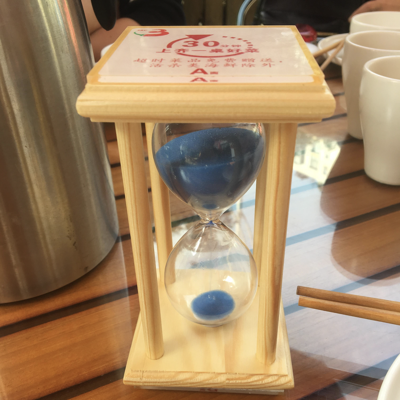 A wooden hourglass with blue sand sits on a table with cups, chopsticks, and a metal container. Chinese text is visible on a card atop the hourglass.