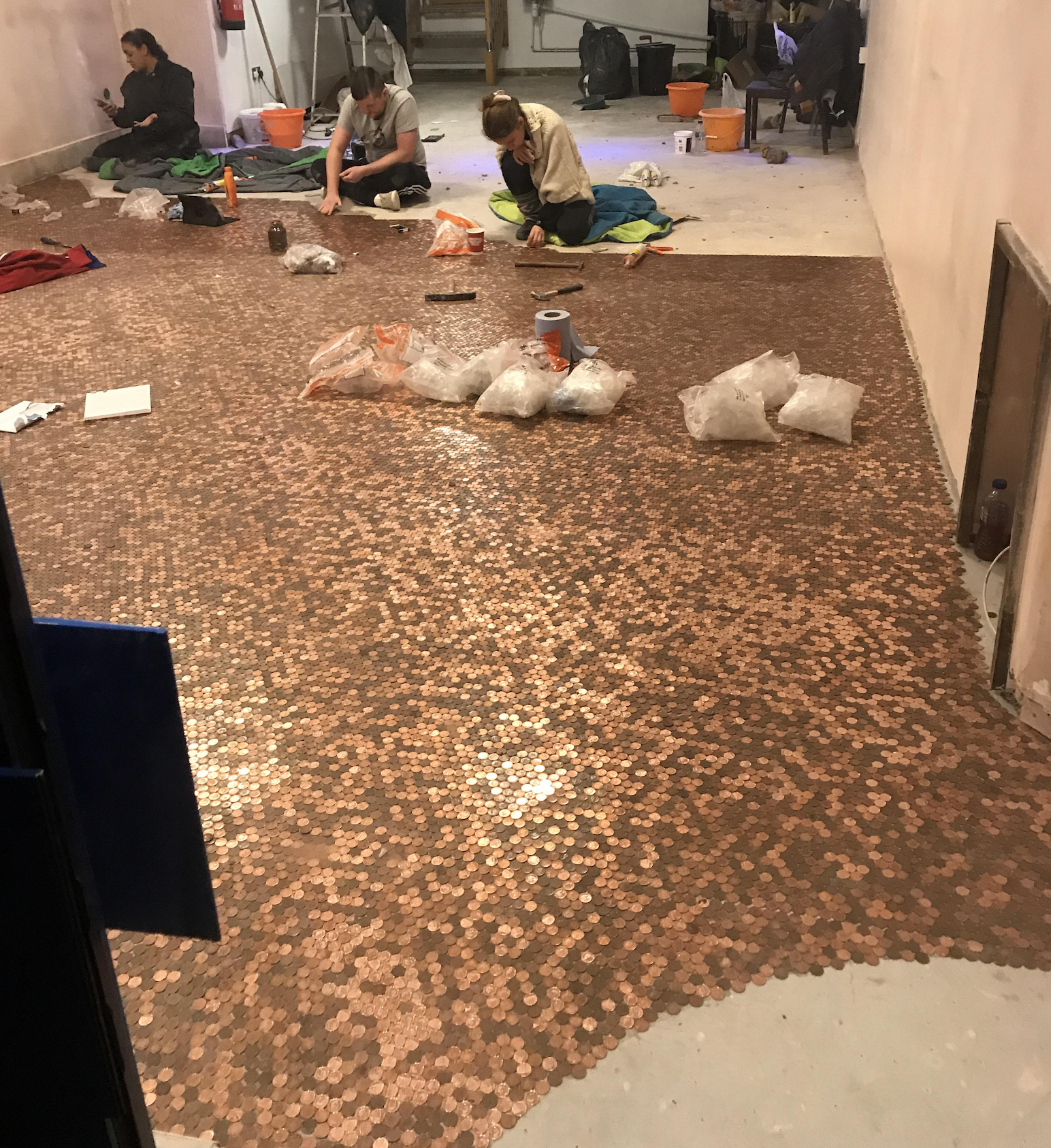 Three people are seen gluing and arranging pennies to cover a floor, creating a mosaic pattern. Buckets, bags of coins, and tools are scattered around the unfinished area.