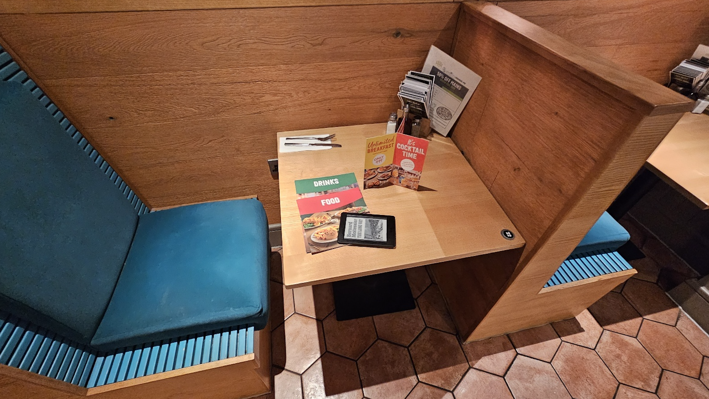 A wooden booth with teal seats, a table set for two, colorful menus, a Kindle, napkins, and utensils, all on a tiled floor in a restaurant.