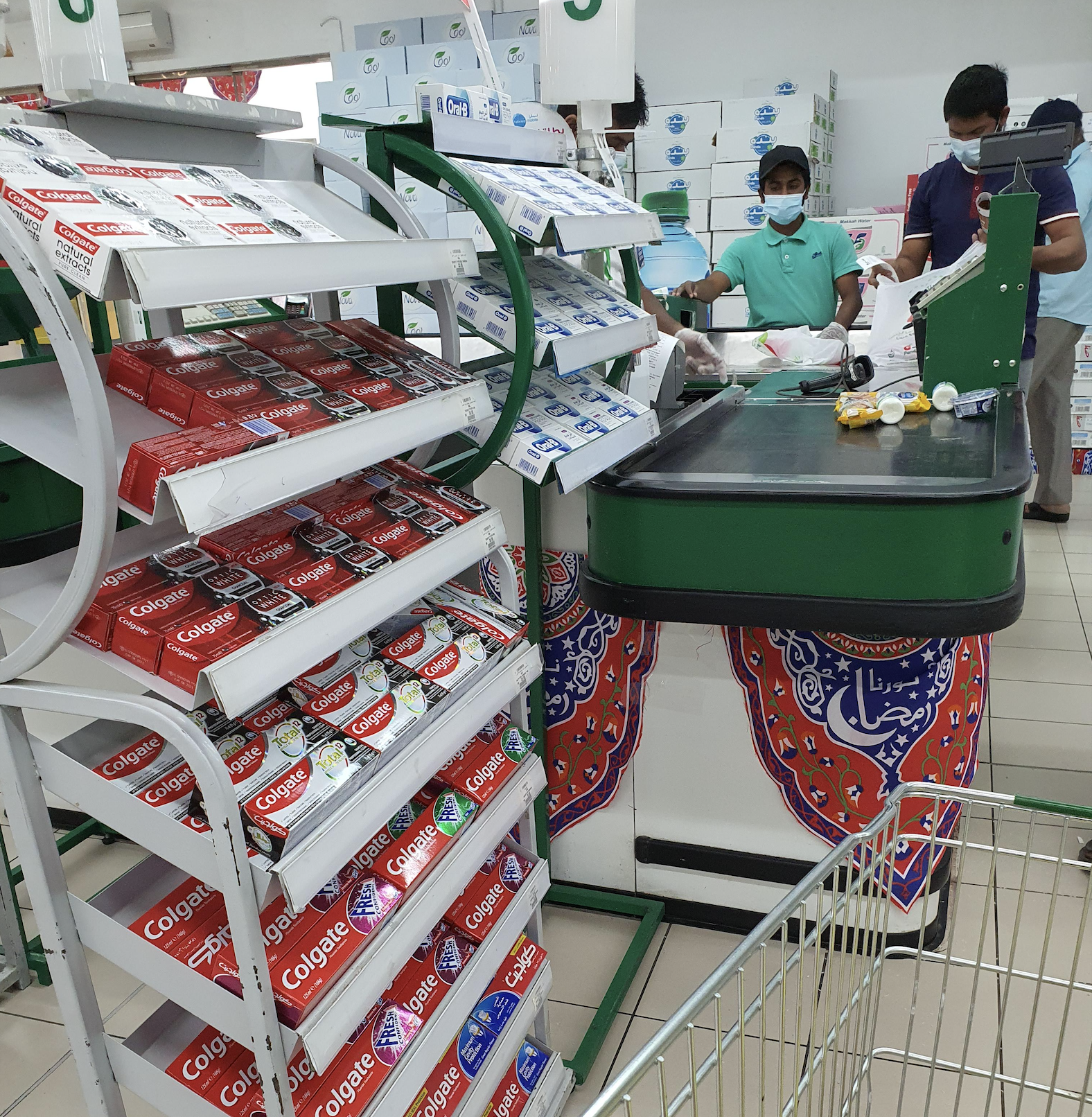 Supermarket checkout area with toothpaste on display, two people wearing masks at the counter, and a shopping cart in the foreground. Various products are on the conveyor belt and shelves in the background.