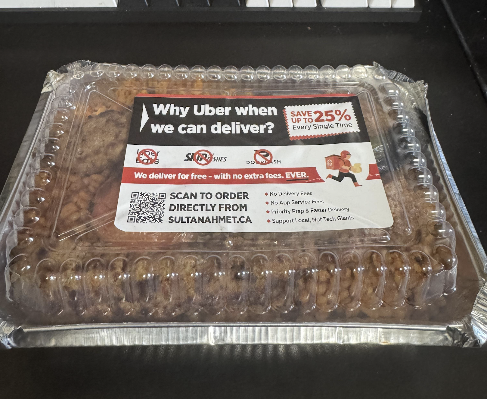 A clear takeout container with food inside has a label promoting direct restaurant delivery over Uber, highlighting savings, no delivery fees, and a QR code to order from sultanahmet.ca.