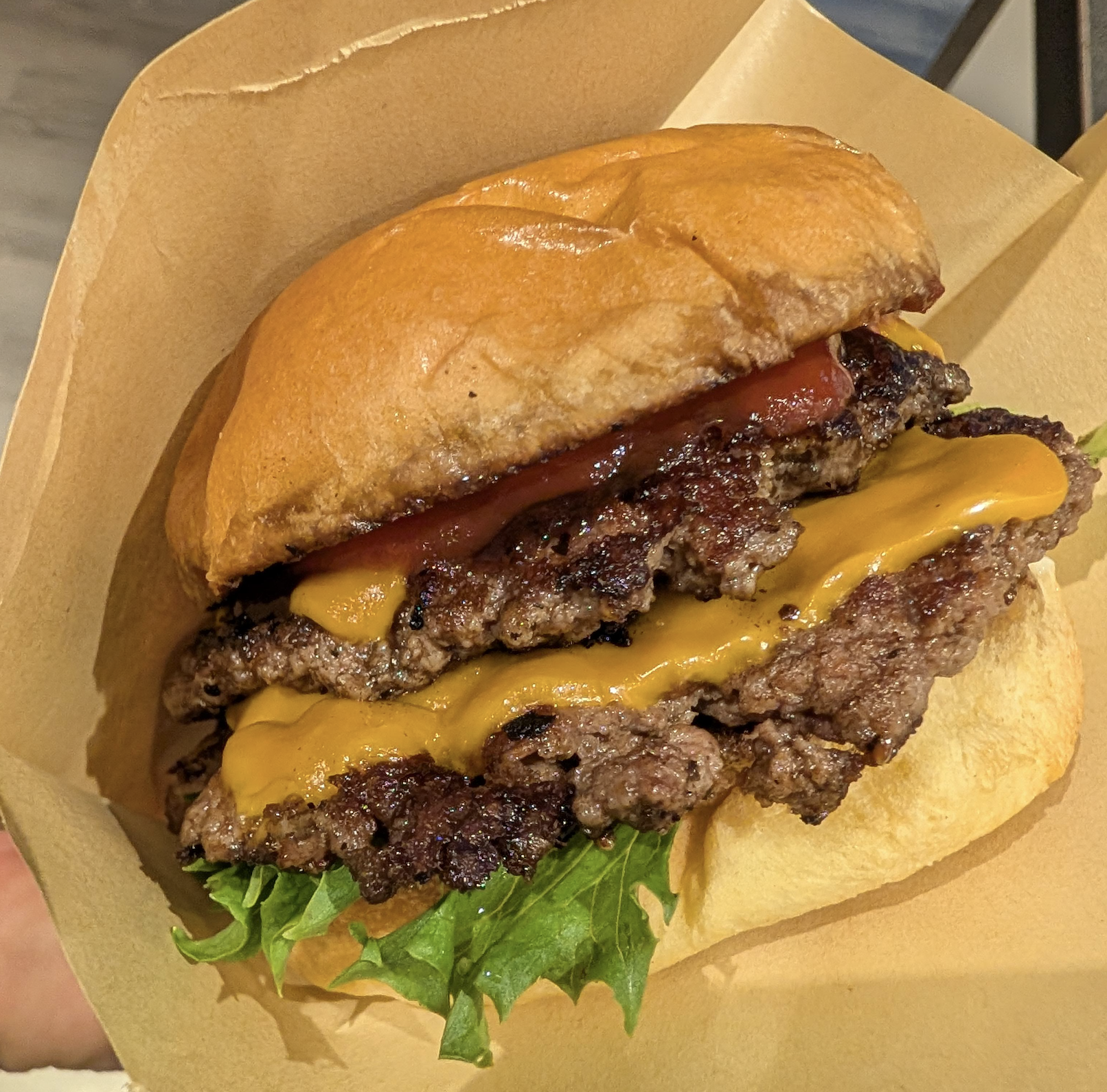 A cheeseburger with two beef patties, melted cheddar cheese, lettuce, and ketchup in a soft bun, served in a brown paper tray.