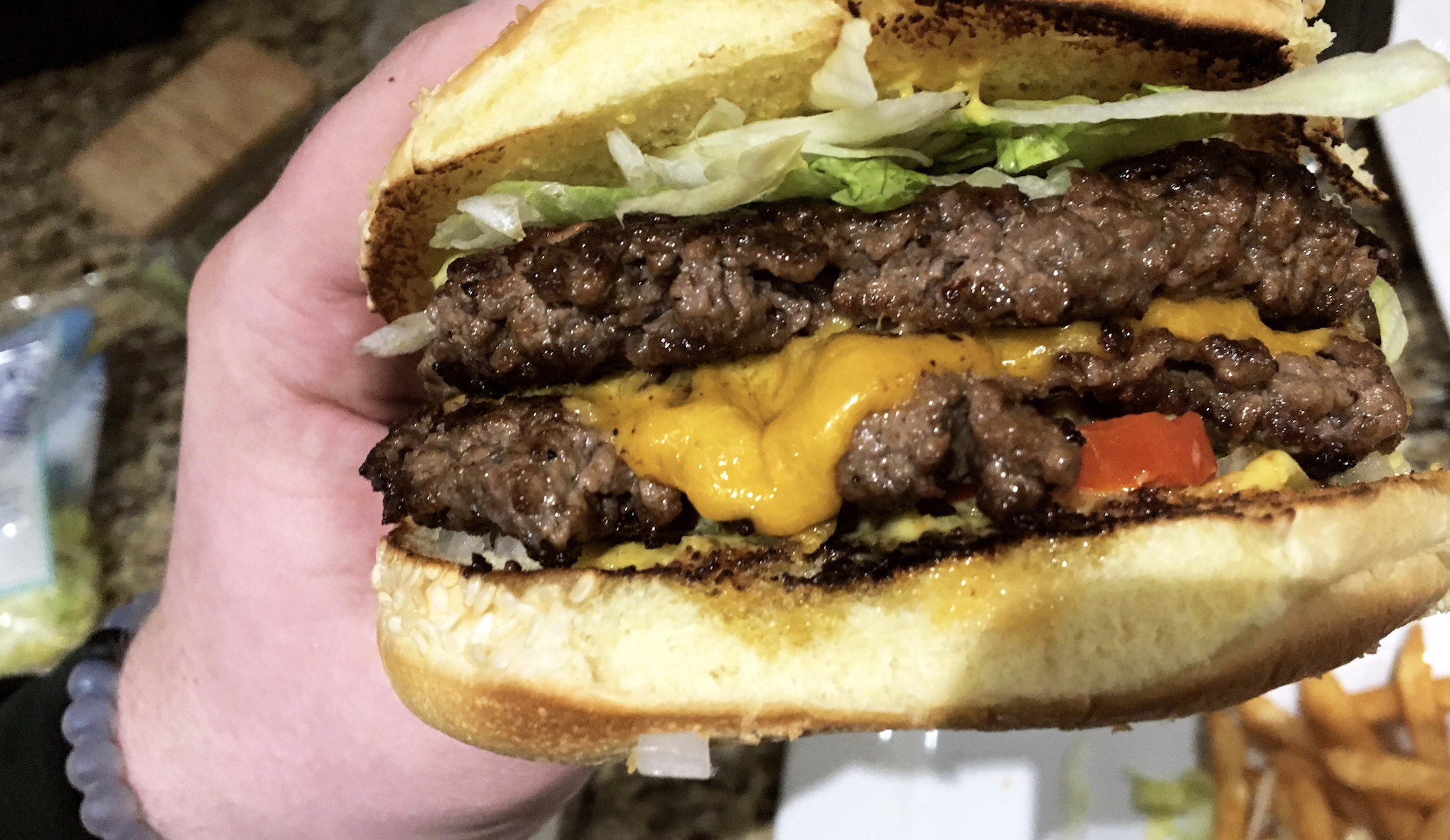A hand holds a cheeseburger with two beef patties, melted cheddar cheese, lettuce, tomato, and a toasted bun. Fries are visible in the background.
