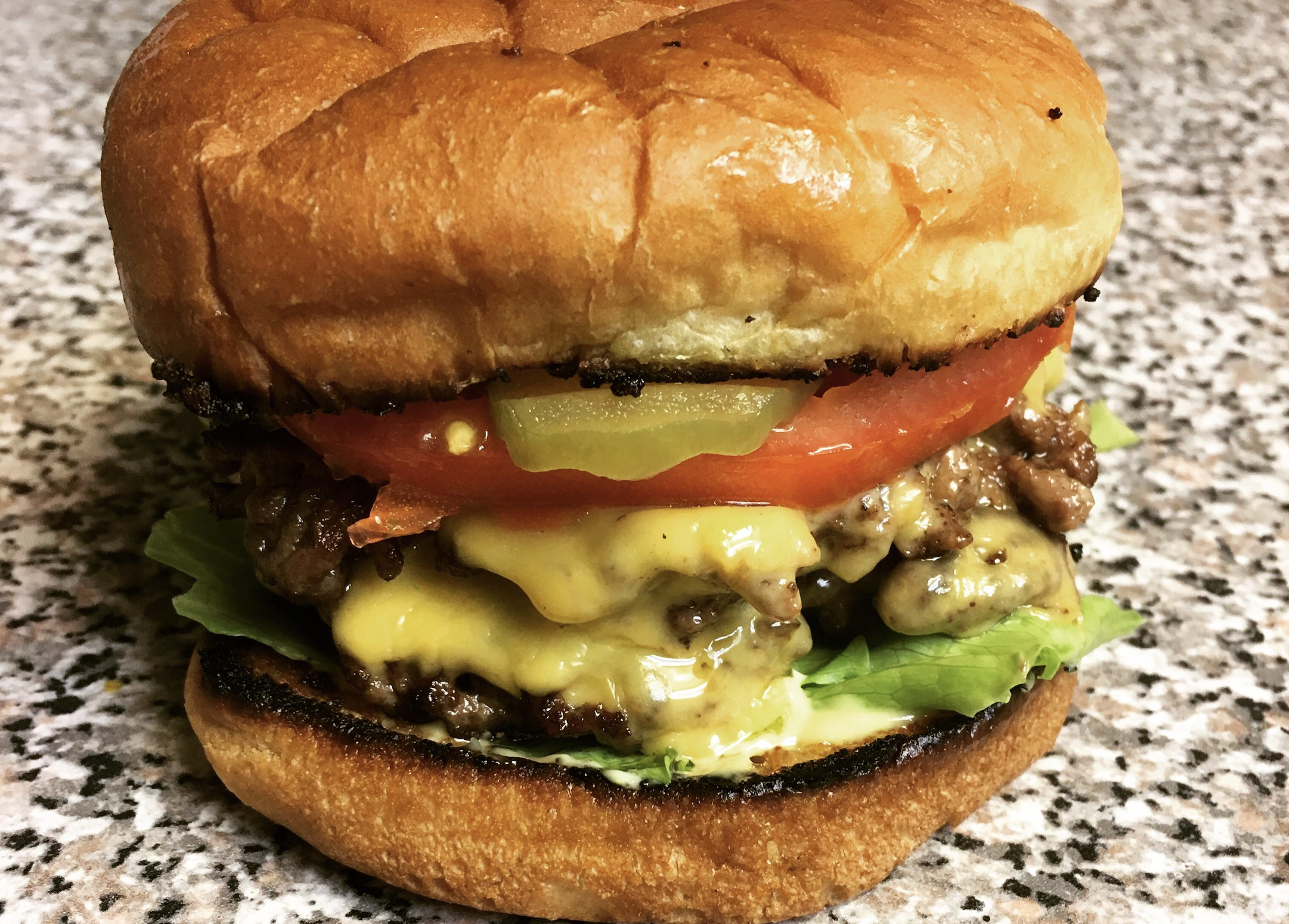 A cheeseburger with lettuce, tomato, pickles, melted cheese, and two beef patties on a toasted bun, sitting on a speckled countertop.