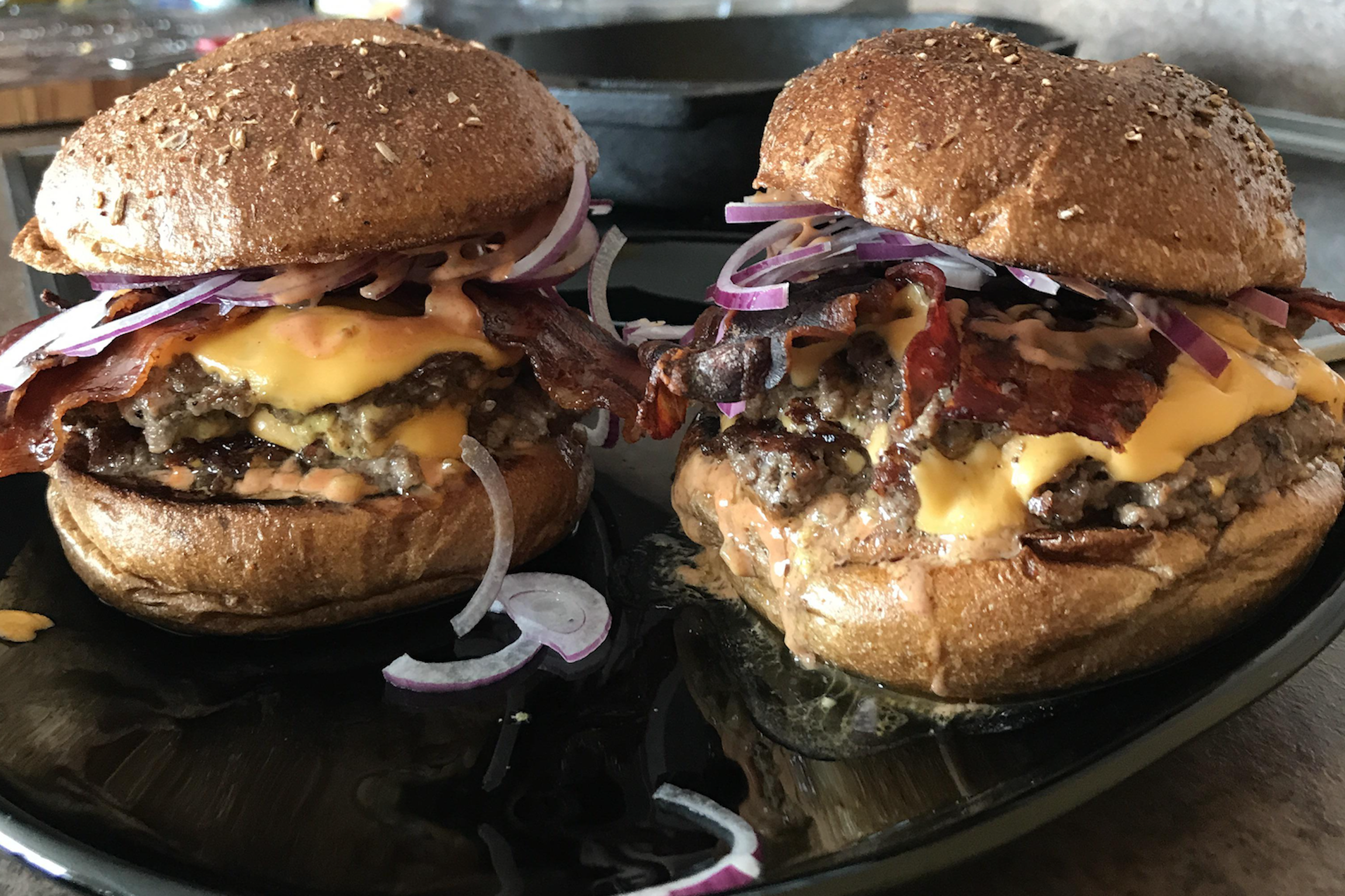 Two cheeseburgers with crispy bacon, melted cheddar cheese, sliced red onions, and a special sauce on toasted whole wheat buns, served on a black plate.