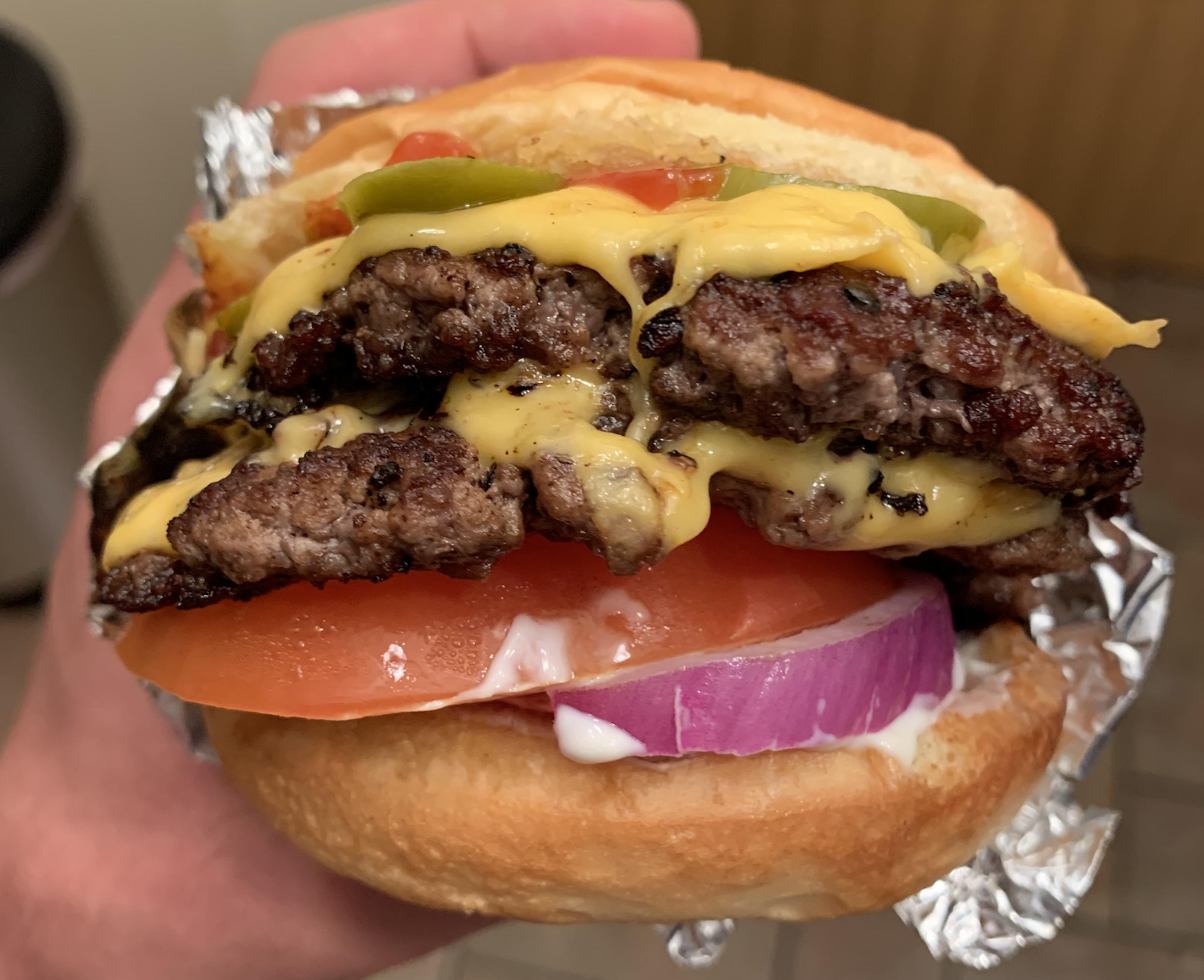 A hand holds a cheeseburger with two beef patties, melted cheese, ketchup, pickles, tomato, red onion, and mayonnaise, wrapped partly in foil.