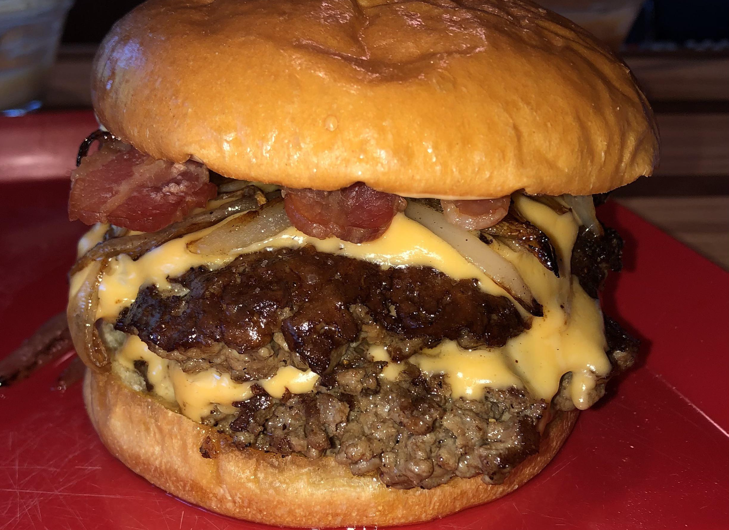 Close-up of a cheeseburger with melted cheese, grilled onions, crispy bacon, and a beef patty on a toasted bun, served on a red plate.