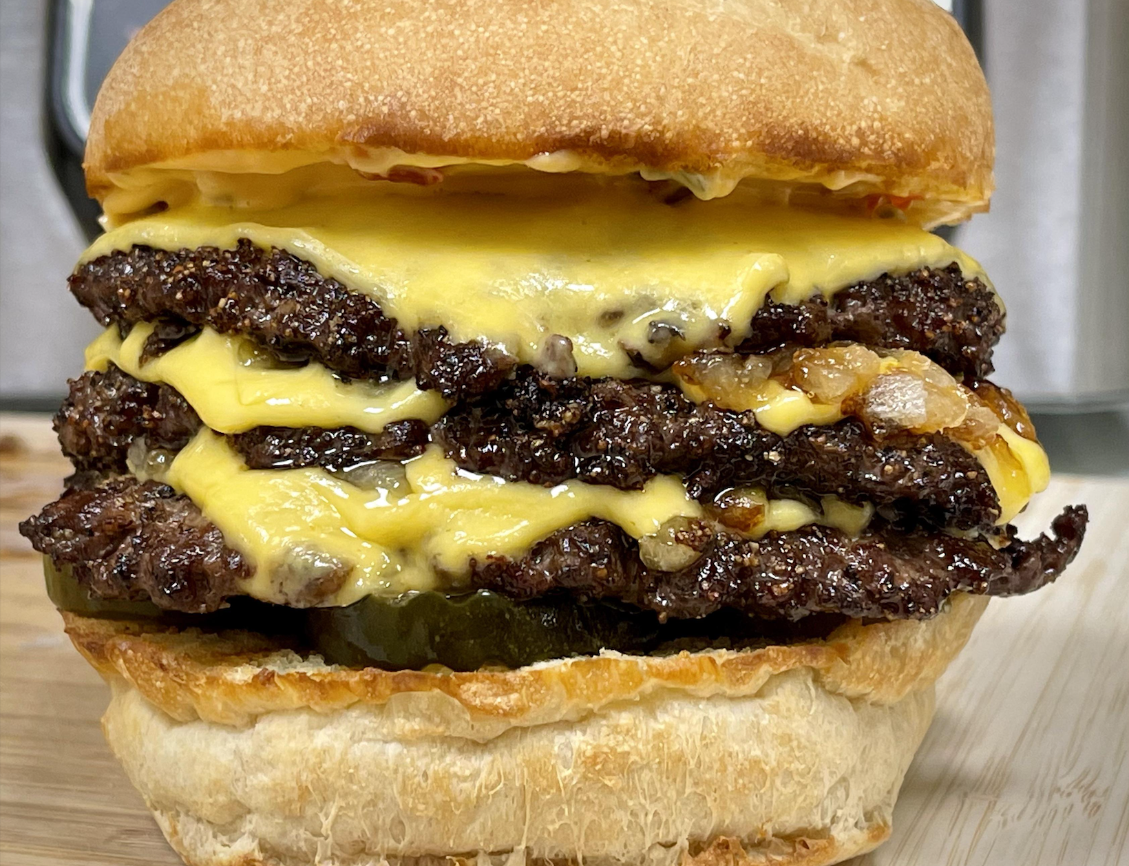 A close-up of a cheeseburger with three beef patties, melted cheddar cheese, pickles, and a toasted bun, set on a wooden surface.