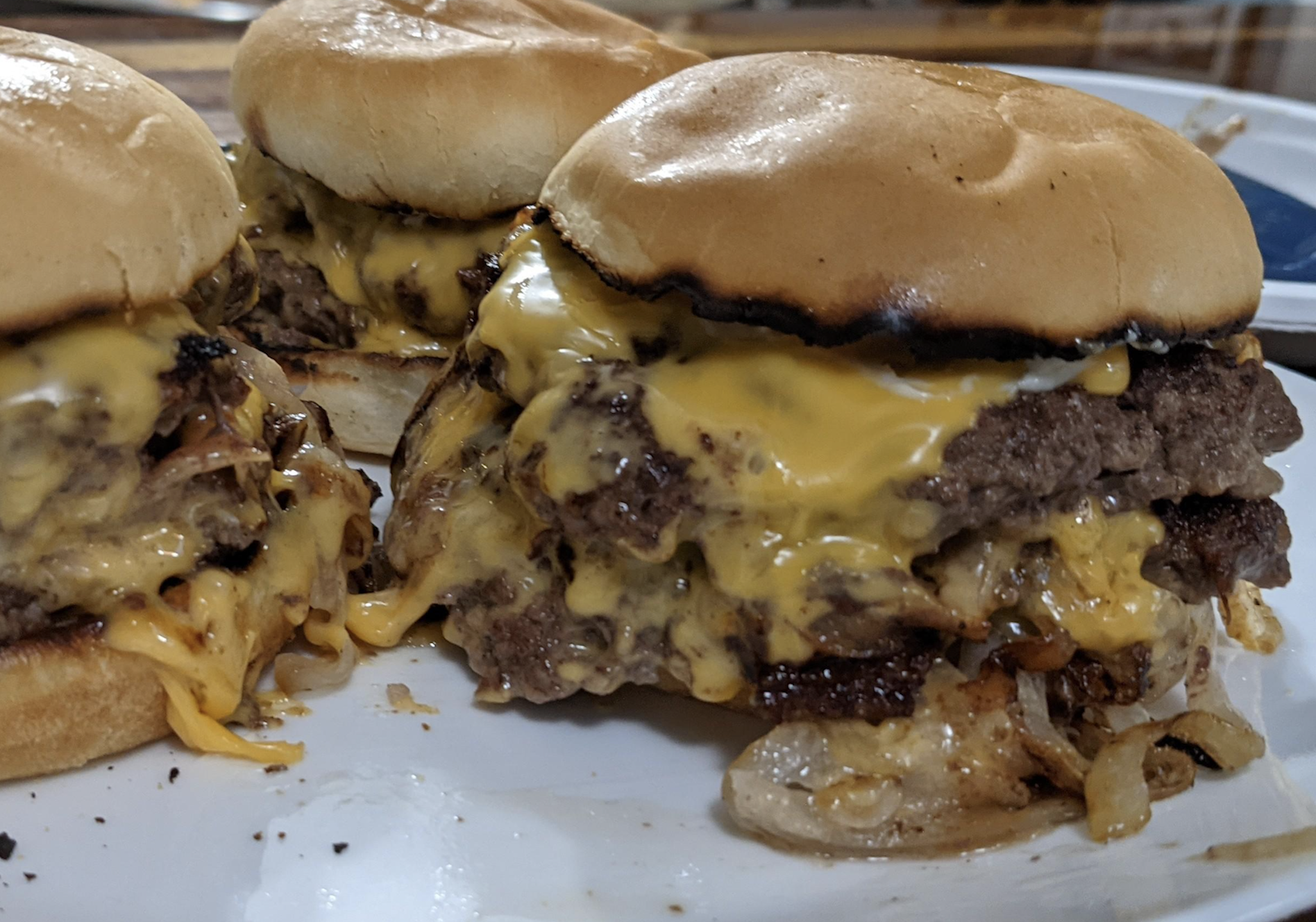 Three cheeseburgers with melted cheese, grilled onions, and beef patties on toasted buns, arranged on a white plate with some cheese and onions spilling out.