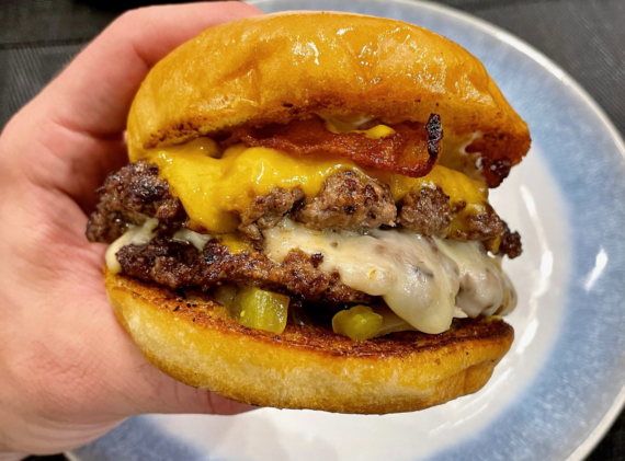A hand holds a cheeseburger with two beef patties, melted cheddar and Swiss cheese, bacon, pickles, and a toasted bun, shown on a plate in the background.