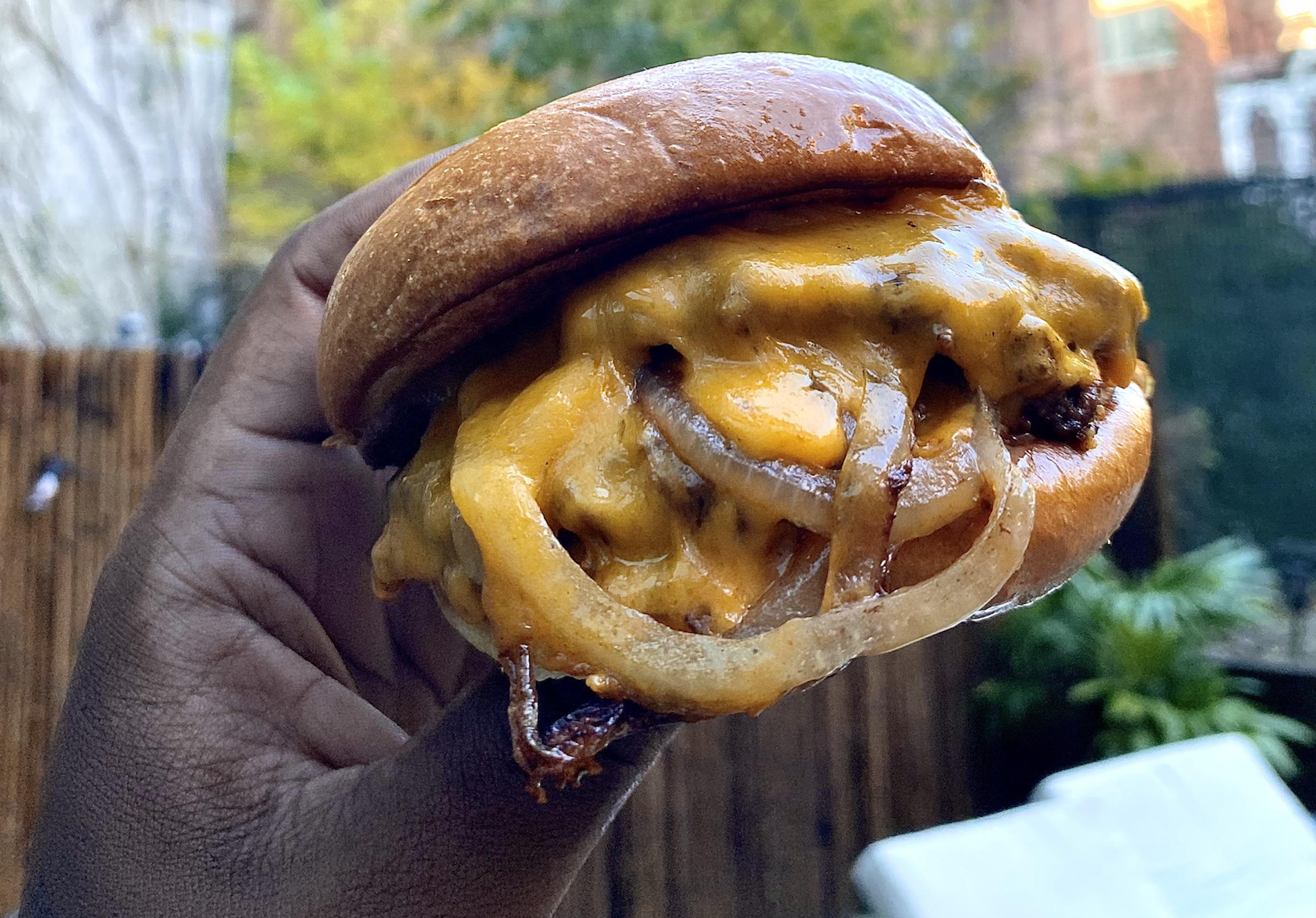 A hand holds a cheeseburger with melted cheddar cheese, grilled onions, and a soft, shiny bun. The background is outdoors with greenery and a wooden fence.
