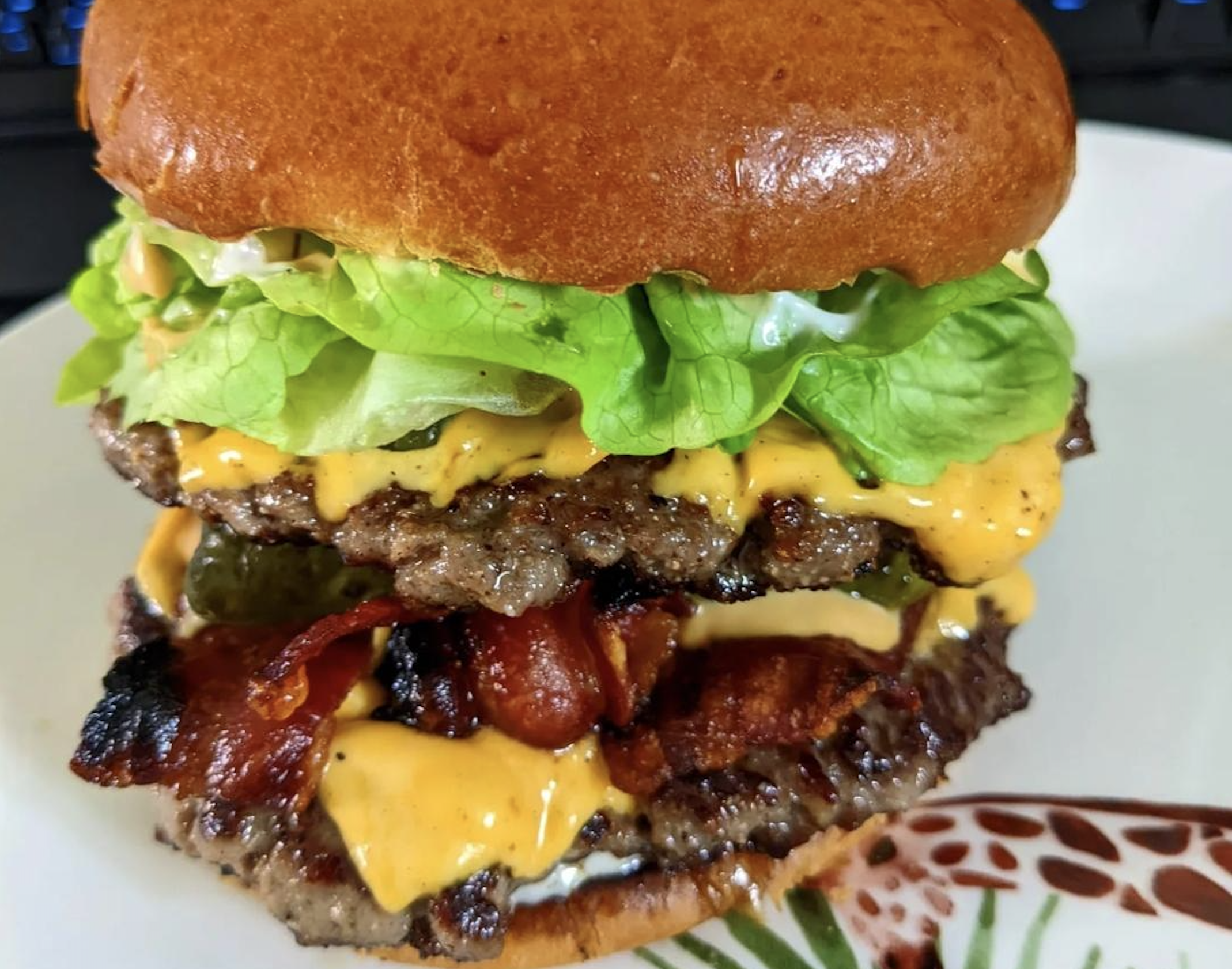 A close-up of a double cheeseburger with lettuce, bacon, melted cheese, pickles, and a toasted bun on a plate.